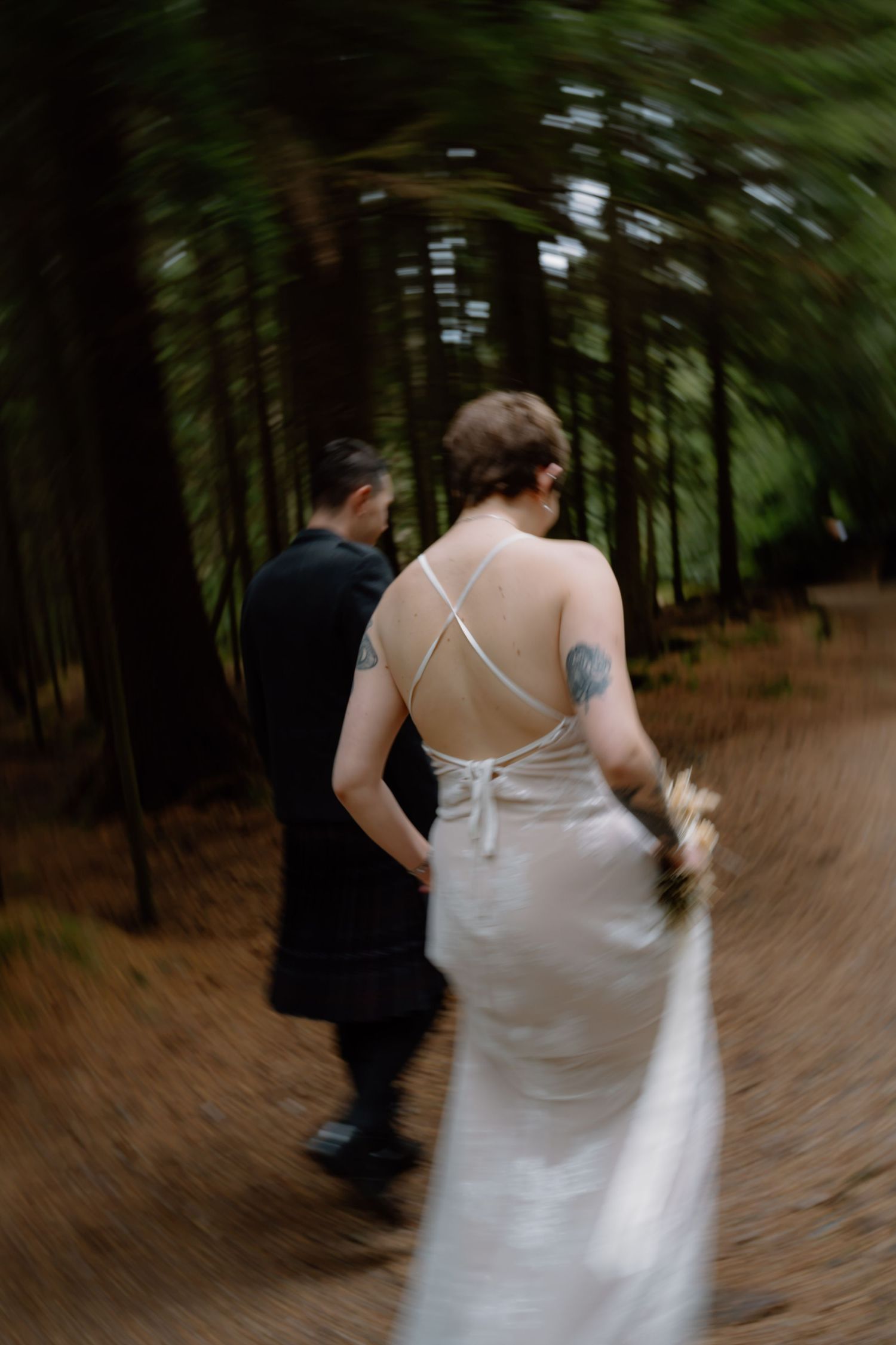 A bride and groom walk through a misty forest path, her backless white gown and tattoos visible in a dreamy blurred shot.