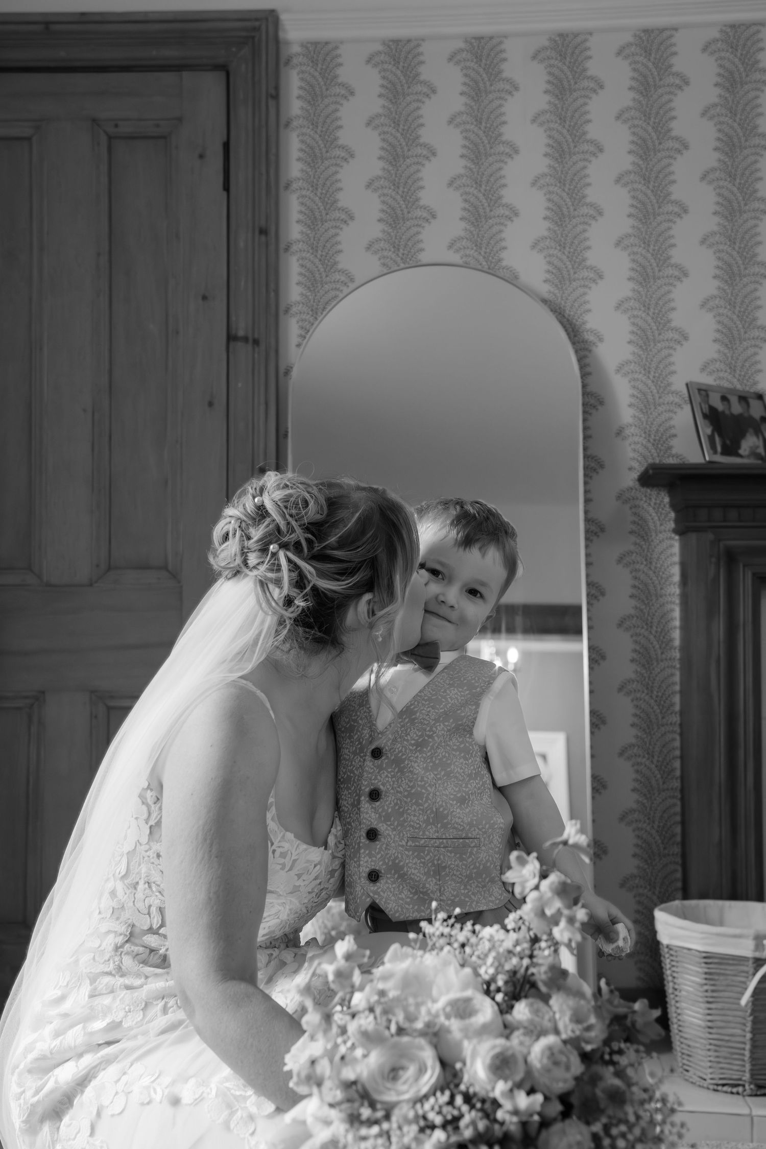 Black and white photo of a bride kissing a young boy in a vest, reflected in an arched mirror, with a floral bouquet.