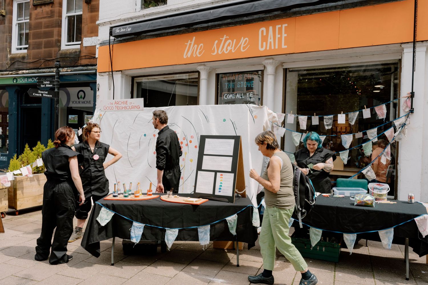 Outdoor market stalls with vendors in black aprons outside The Stove Cafe on a sunny day.