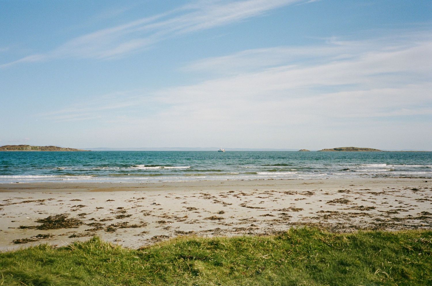 Serene sandy beach with seaweed, turquoise ocean waters, small islands on horizon, and blue sky with wispy clouds.