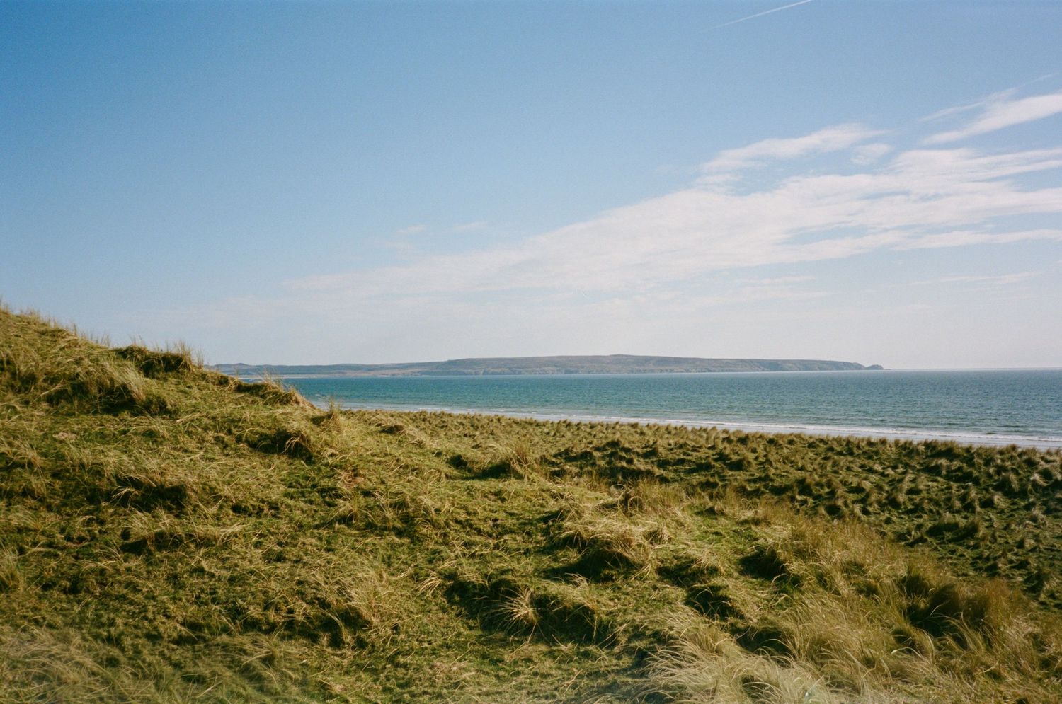 Scenic coastal landscape with grassy sand dunes overlooking a calm blue sea under a clear sky.