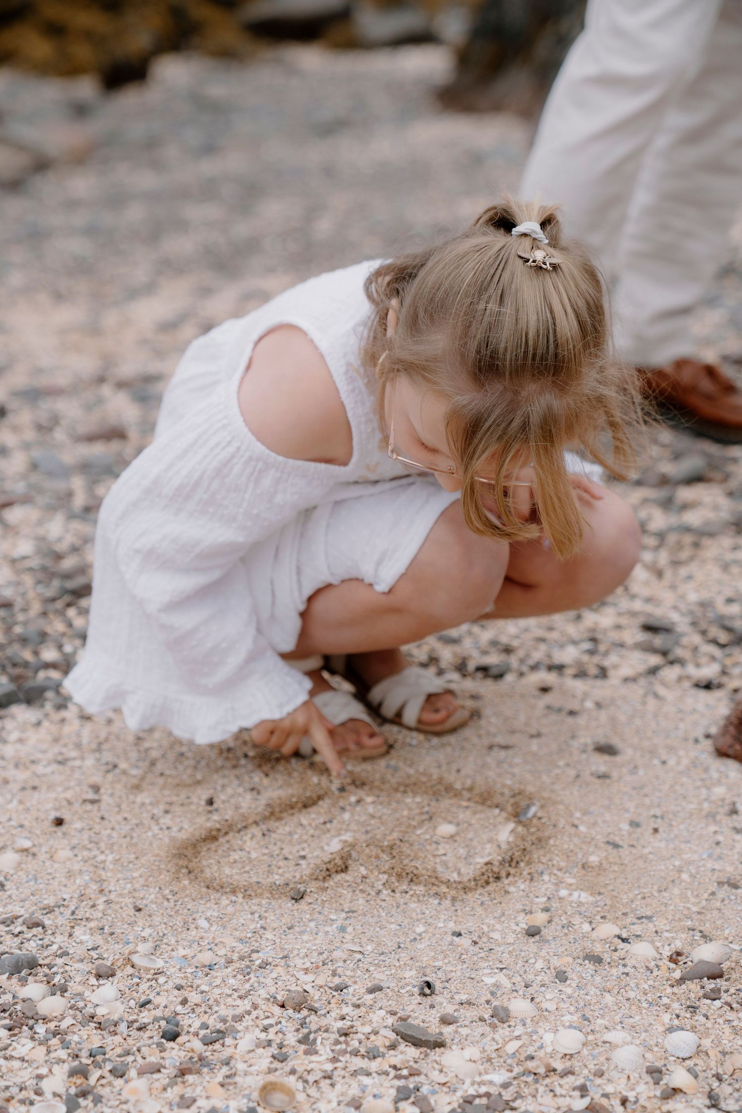 A small child in a white dress explores the sandy beach while crawling.
