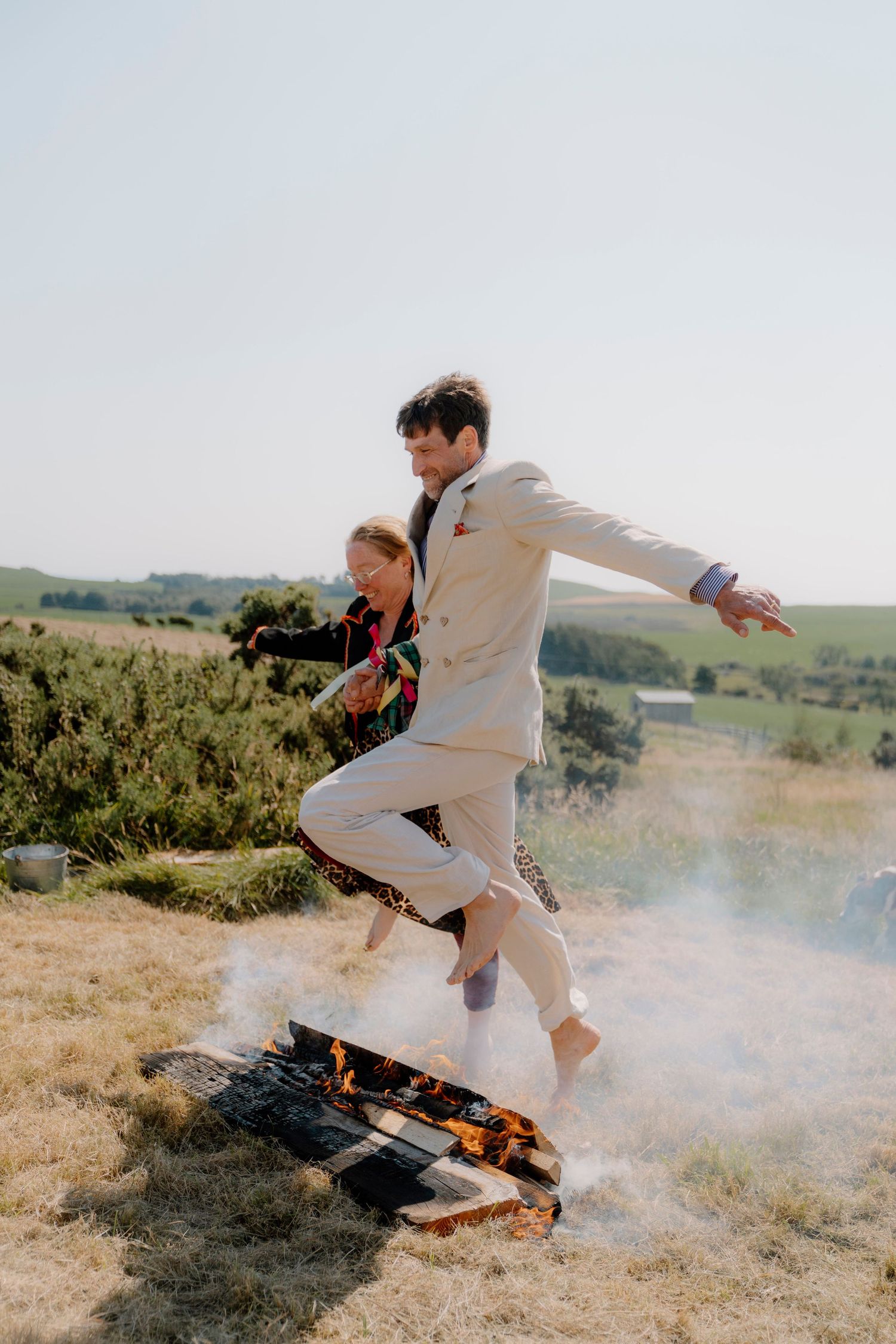 Person in white outfit jumping over bonfire in grassy field.