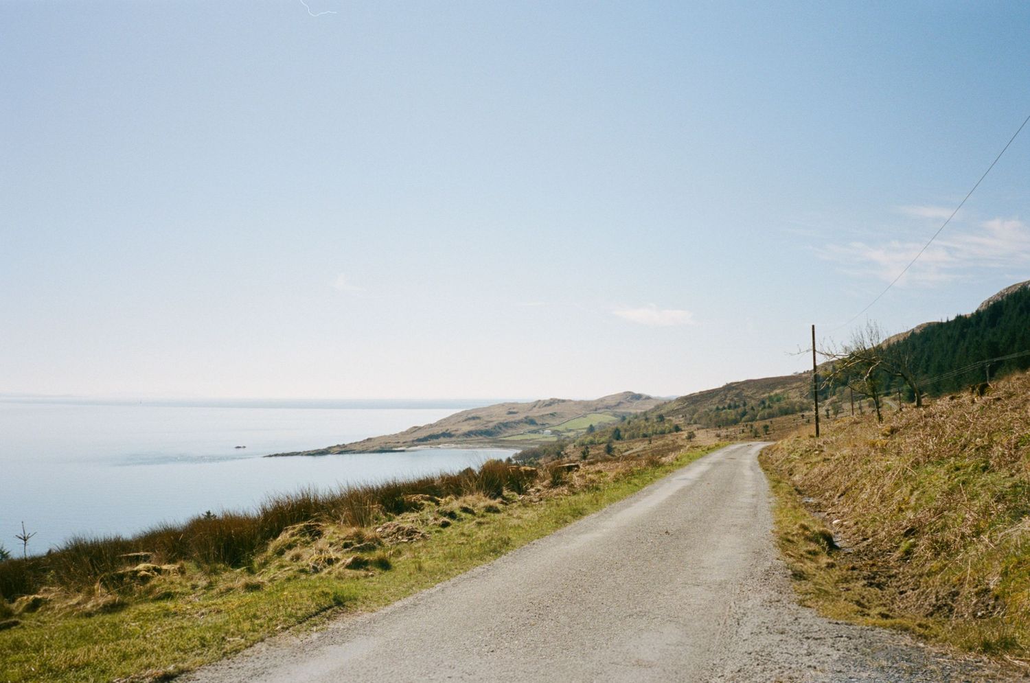 A narrow coastal road winds along green hillsides with stunning ocean views under a clear blue sky.