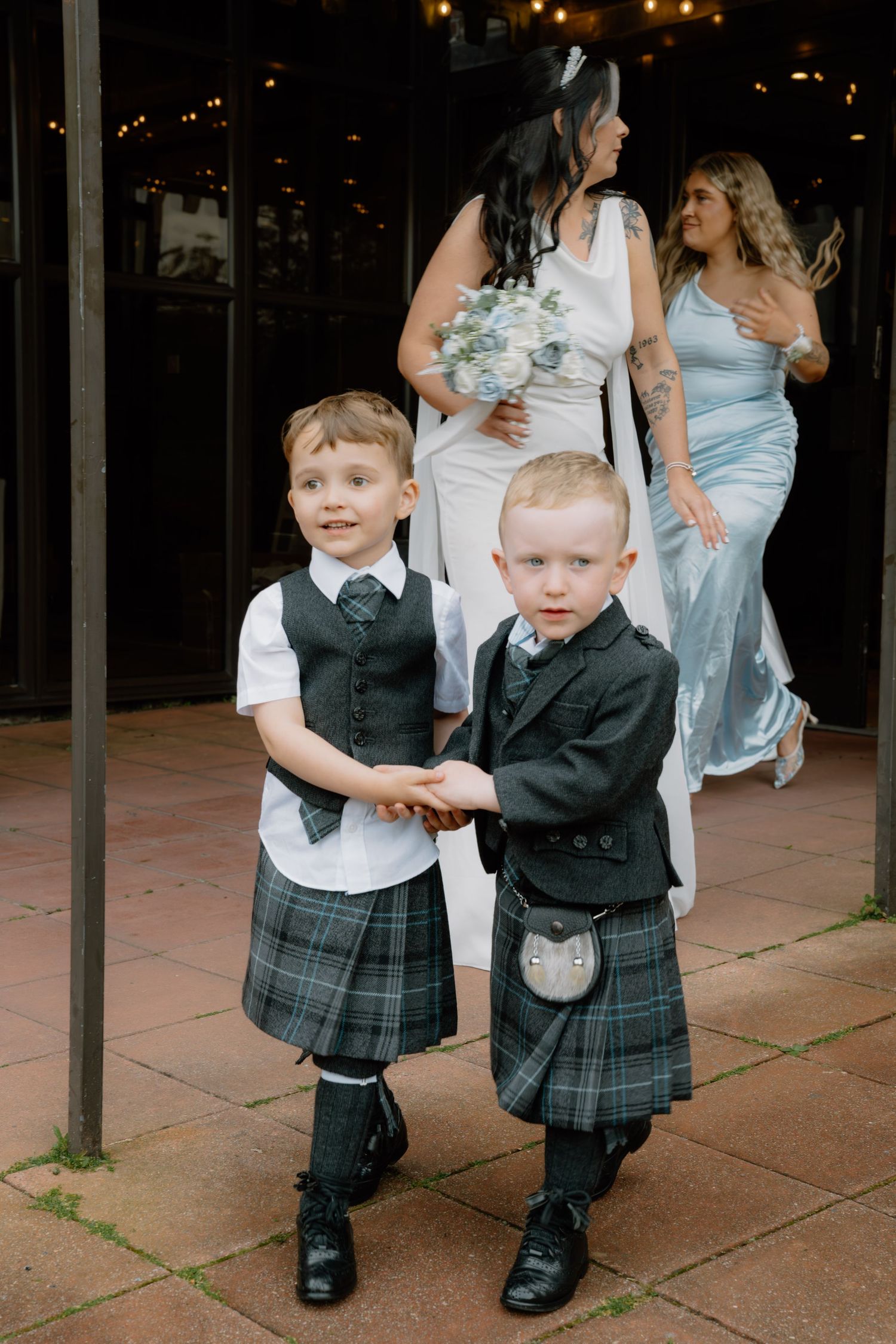 Two young ring bearers in traditional Scottish kilts stand together at a wedding venue.