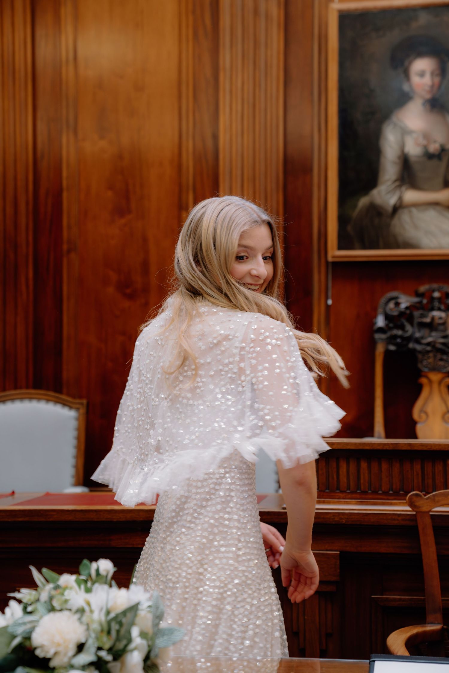 Blonde bride in a pearl-embellished white gown with ruffled sleeves turns to smile in an elegant wood-paneled room.