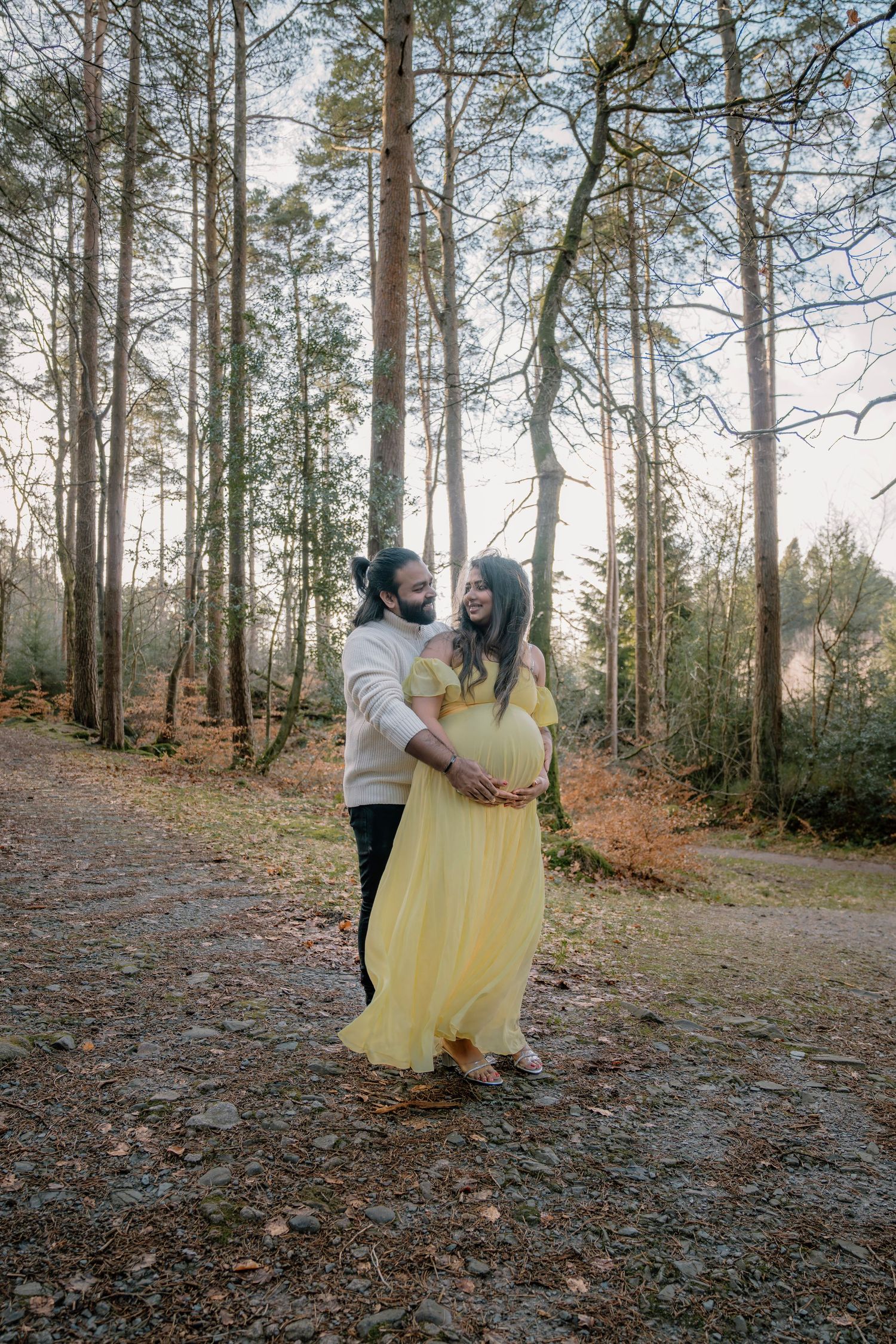 A couple shares a romantic moment on a forest path surrounded by tall pine trees on an autumn day.