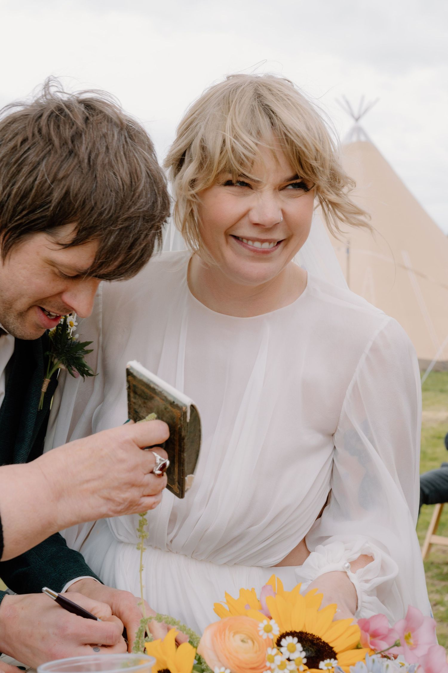 Smiling bride in white dress holds sunflowers while groom signs document at outdoor wedding.