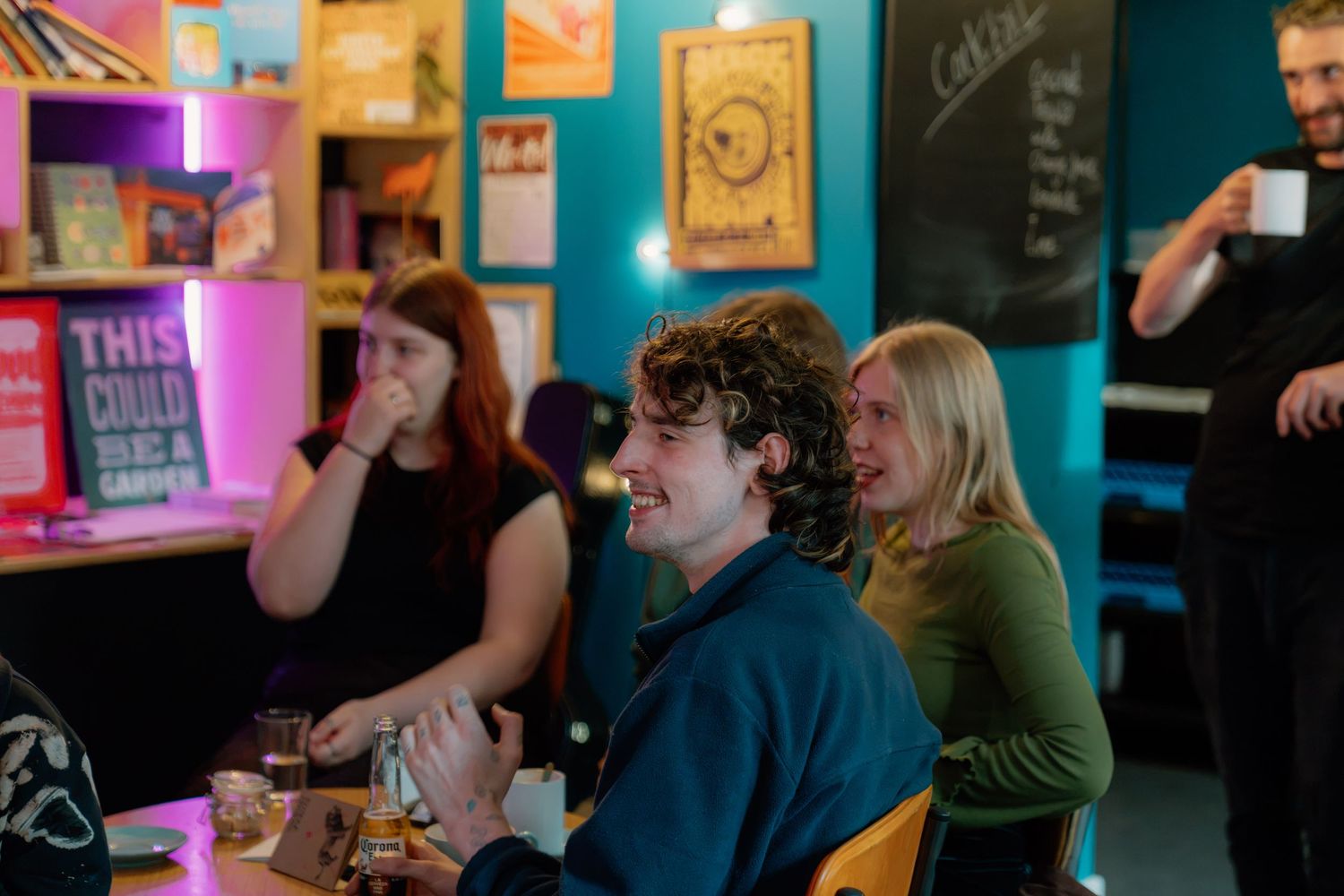 People socializing at a colorful bar with teal walls, pink lighting, and a smiling young man in the foreground.