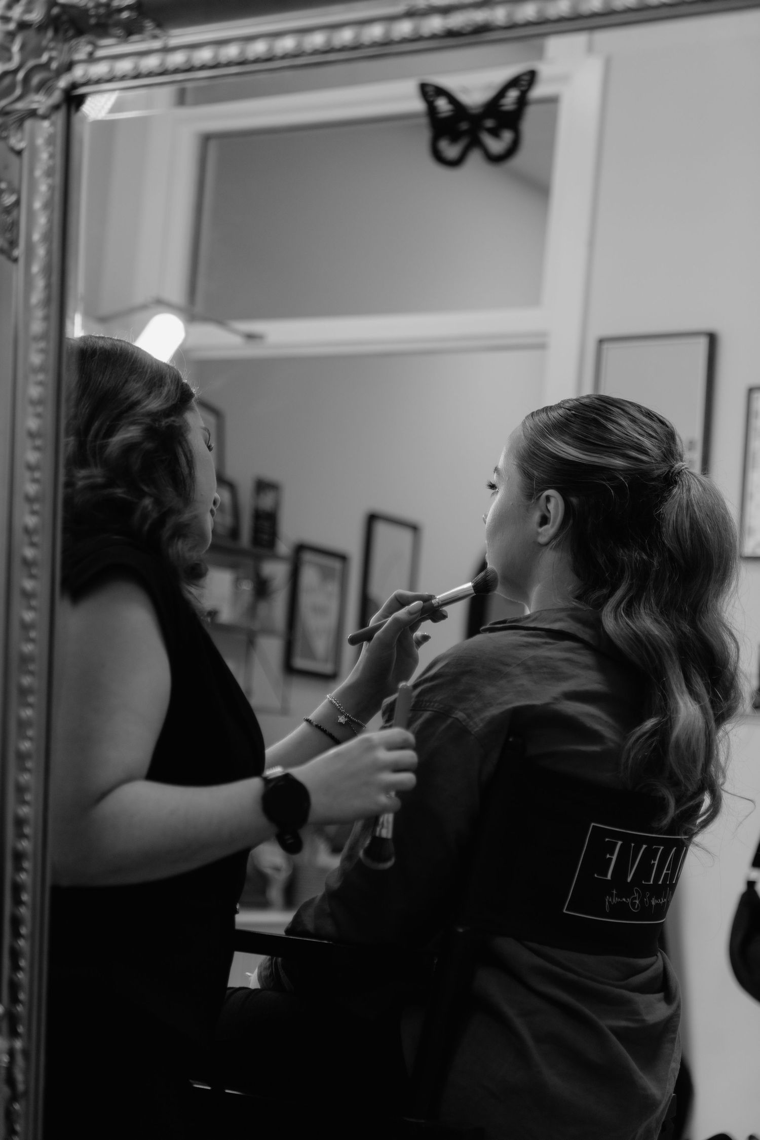 Black and white photo of a makeup artist applying lipstick to a client in a salon mirror.