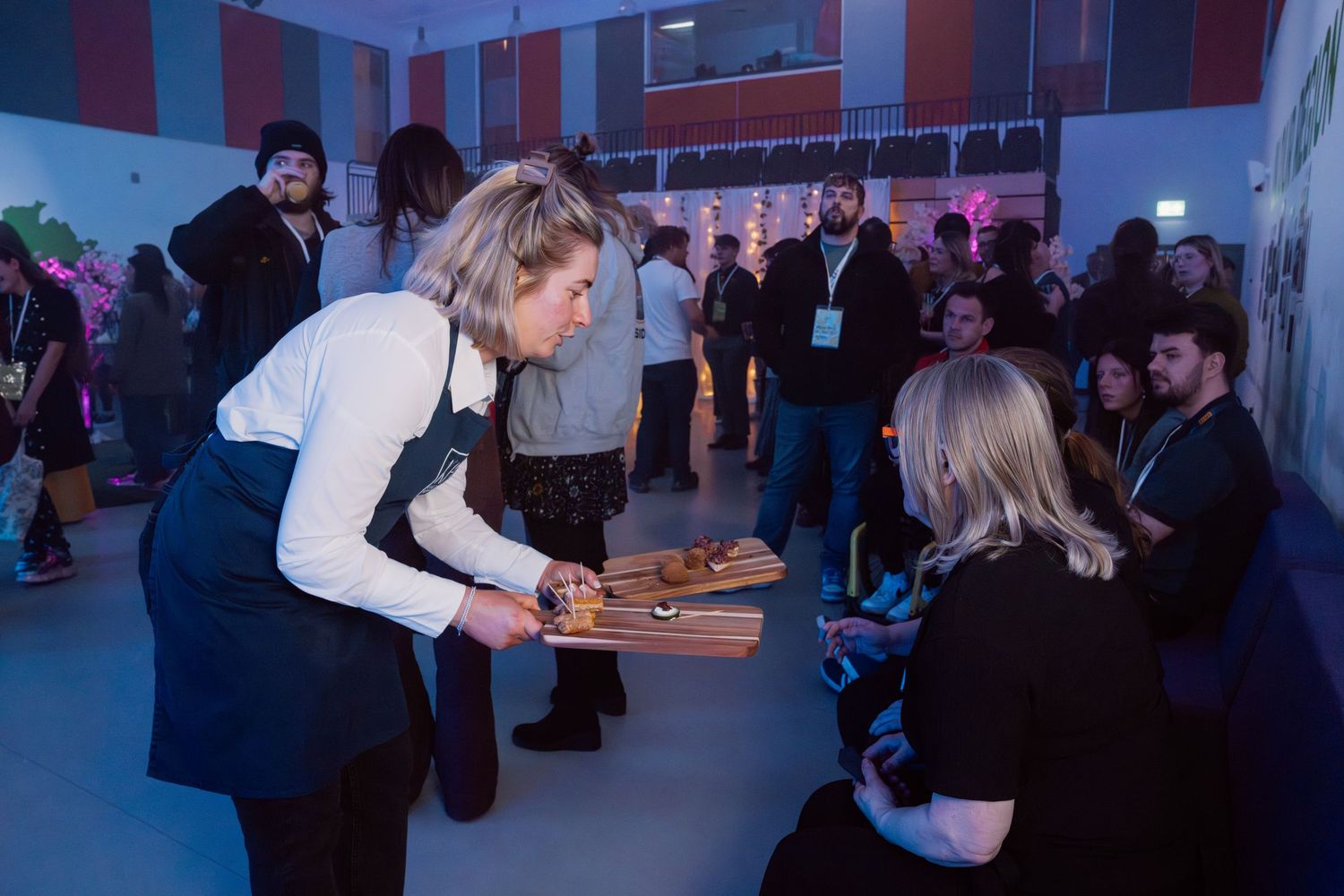 A server offers appetizers on a wooden board to guests at a crowded indoor networking event.