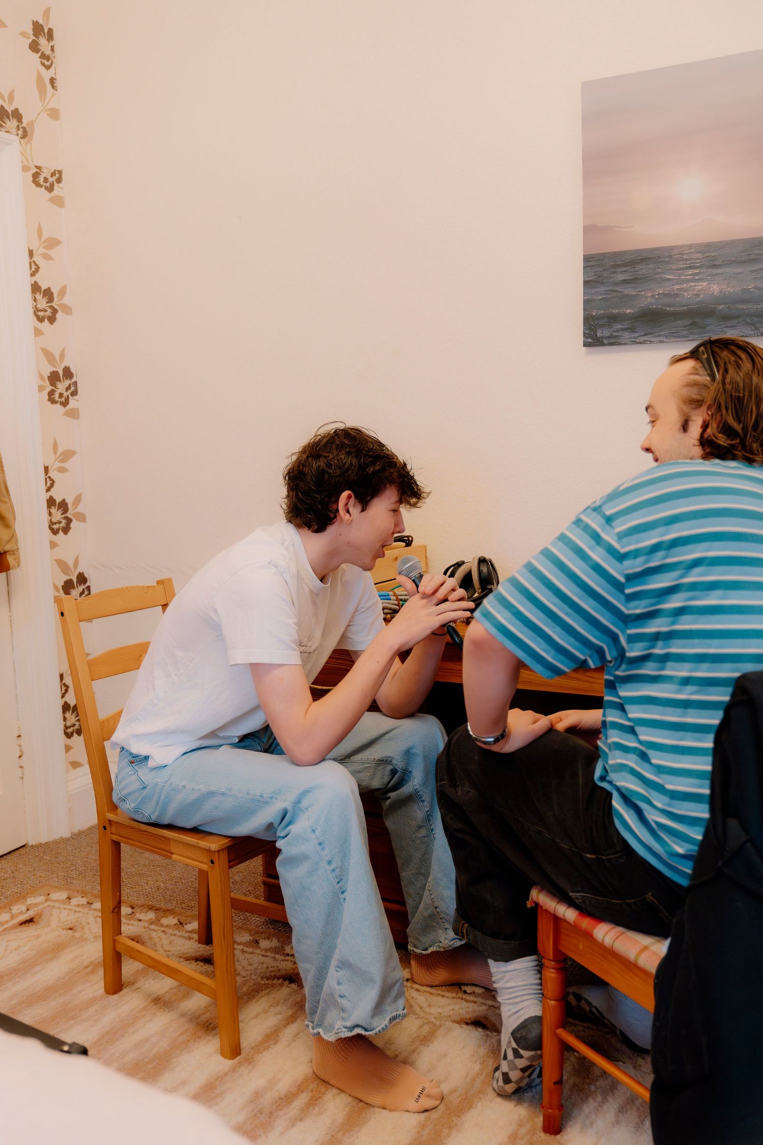 Two young men sit at a wooden table in a cozy room, examining a camera together near floral wallpaper.