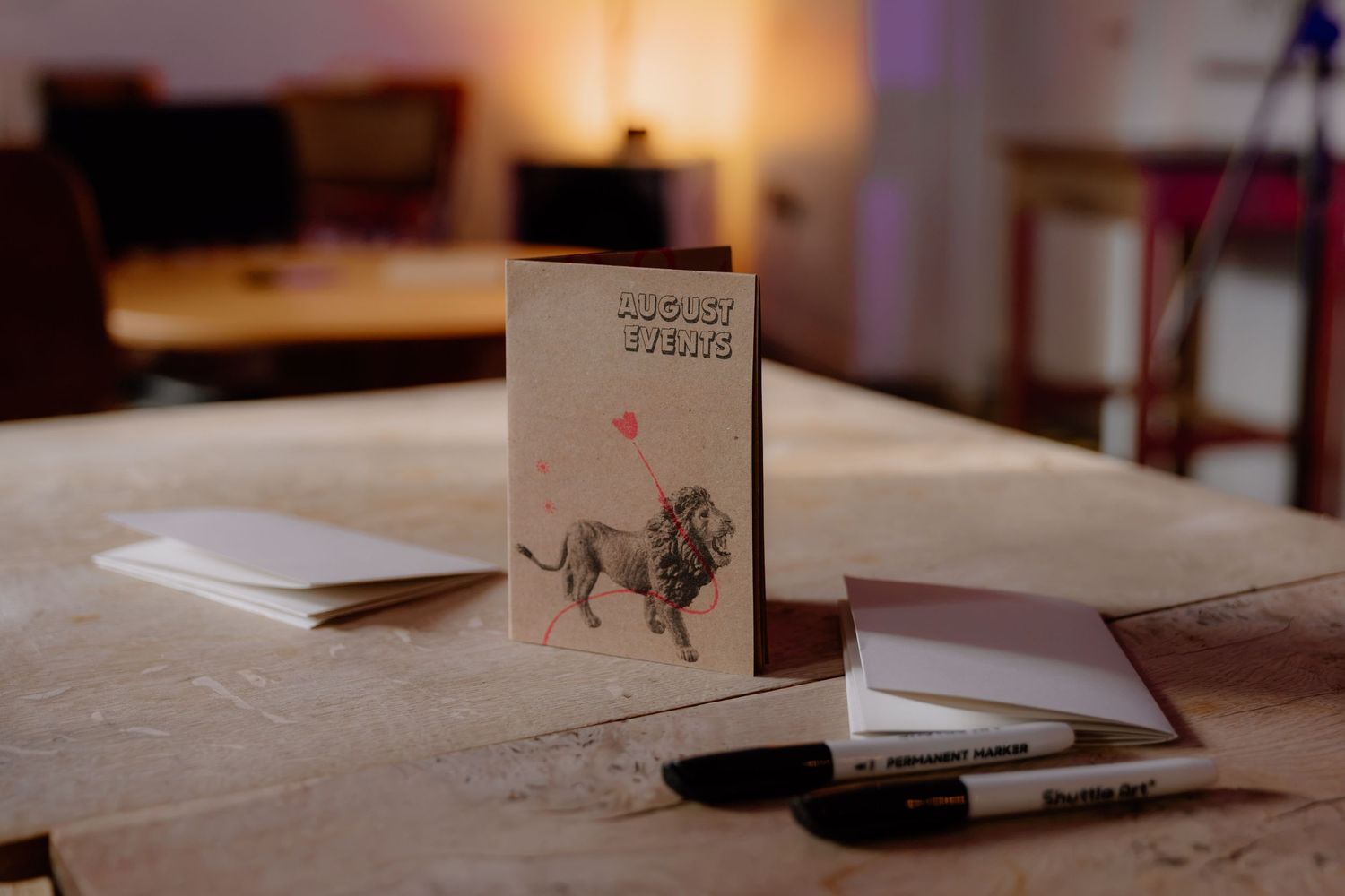 A greeting card featuring a lion illustration stands upright on a wooden table beside markers and notepads.