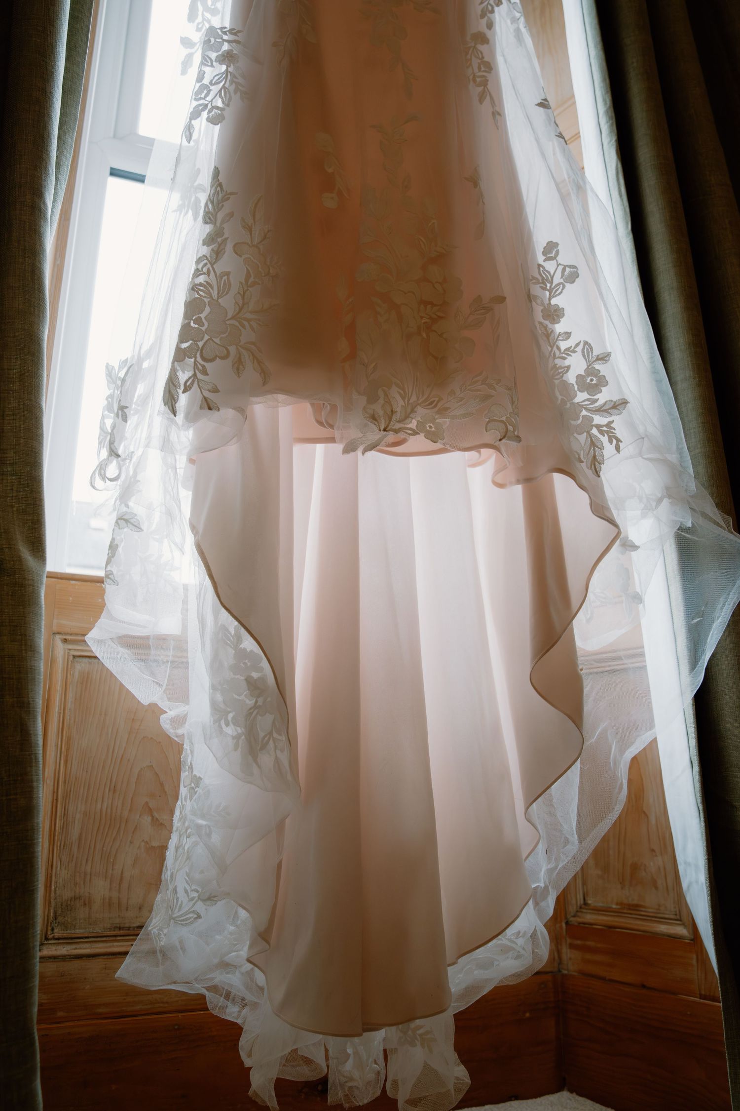 Elegant wedding dress with lace detailing hanging by a sunlit window, showcasing delicate floral embroidery and flowing skirt.