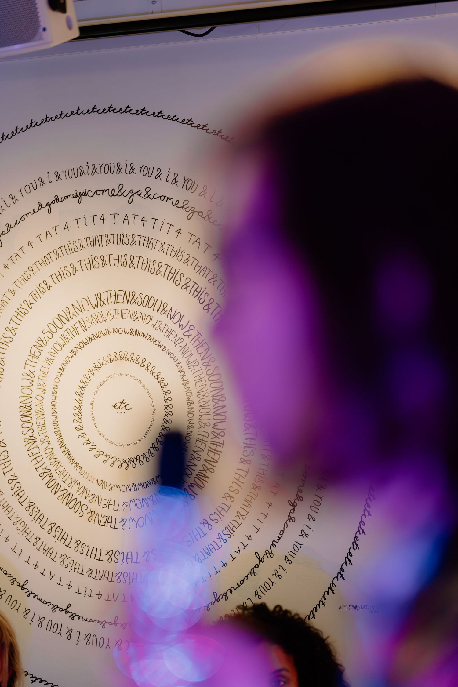 Spiral text art installation on white wall with purple bokeh lights in foreground at an event.