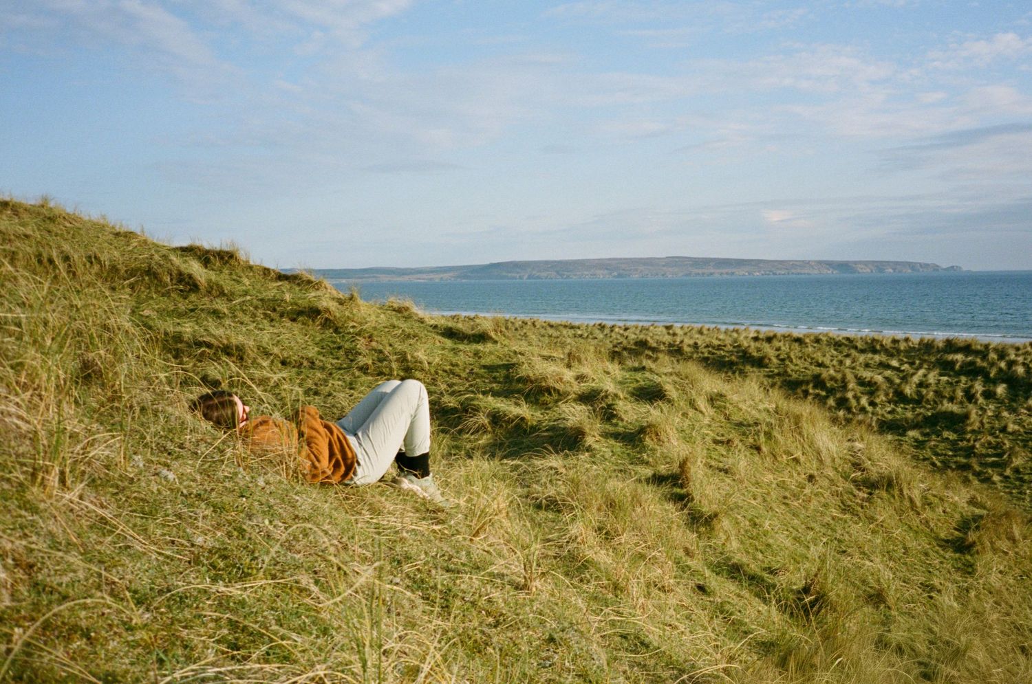 Person relaxing on grassy coastal dunes with scenic ocean and blue sky views in the background.