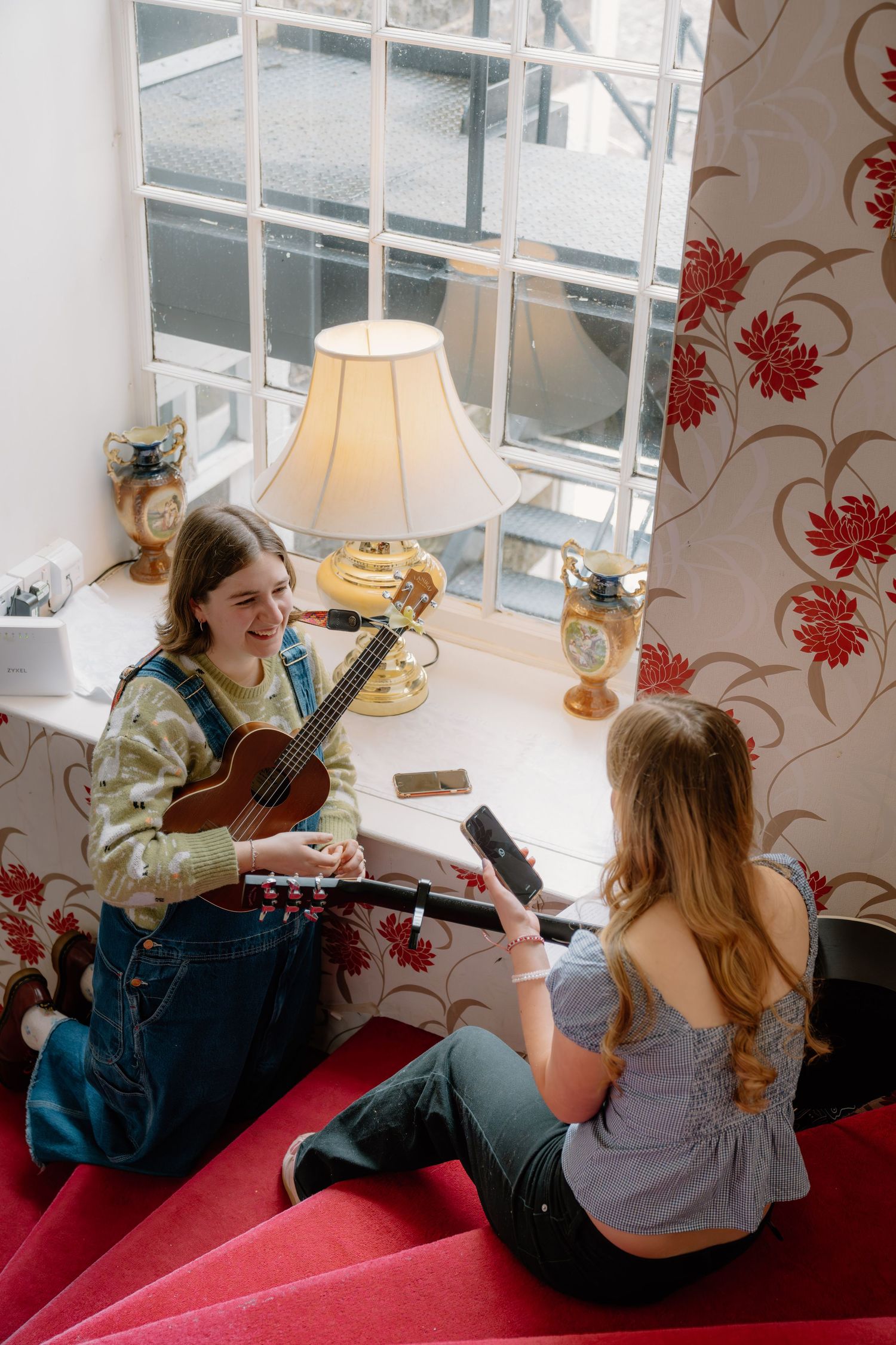 Two teenage girls sitting by a window, one playing ukulele and one holding a book, in a cozy floral wallpapered room.