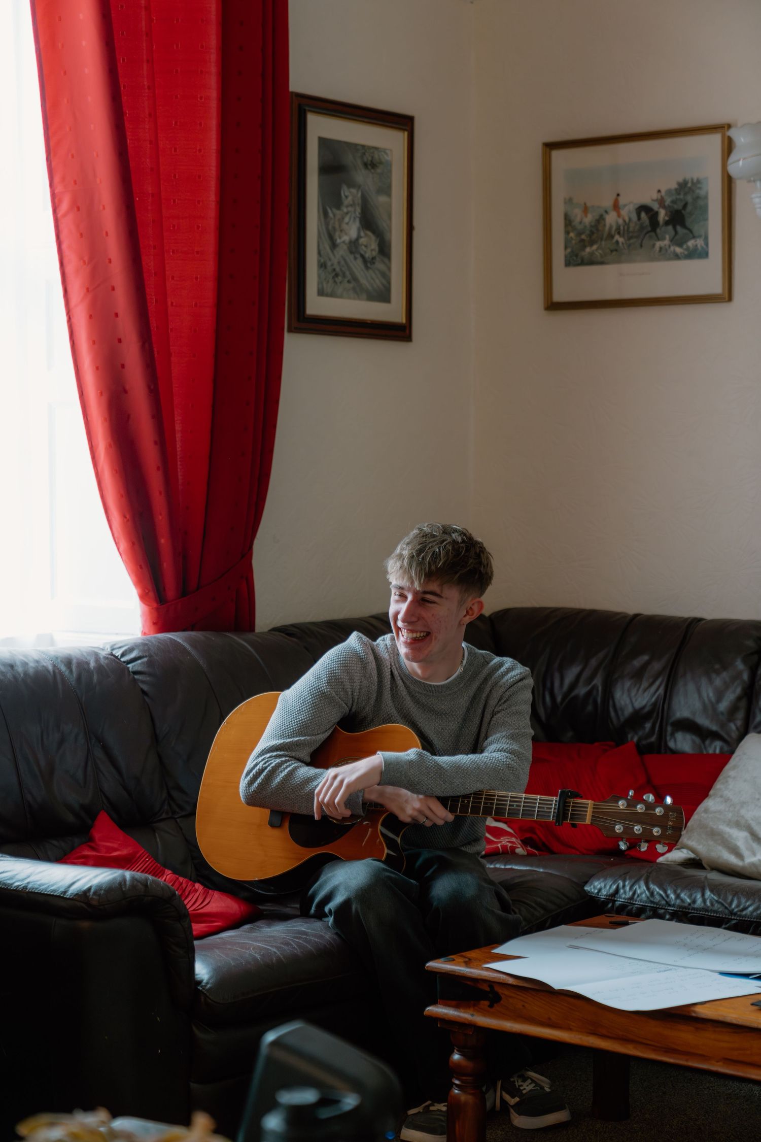 Young man smiling while playing acoustic guitar on a leather sofa in a cozy living room with red curtains.