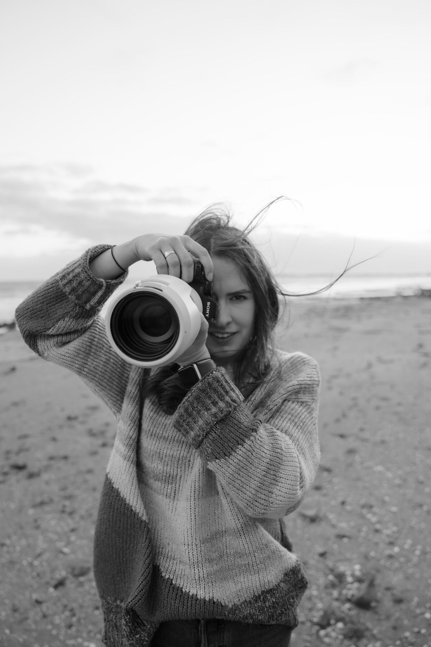 Person in chunky knit sweater takes photo with professional camera on beach.