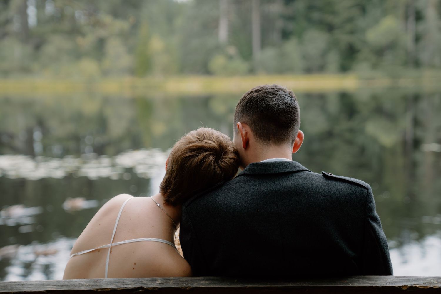 A couple in wedding attire sits on a bench by a tranquil lake, the bride resting her head on the groom's shoulder.