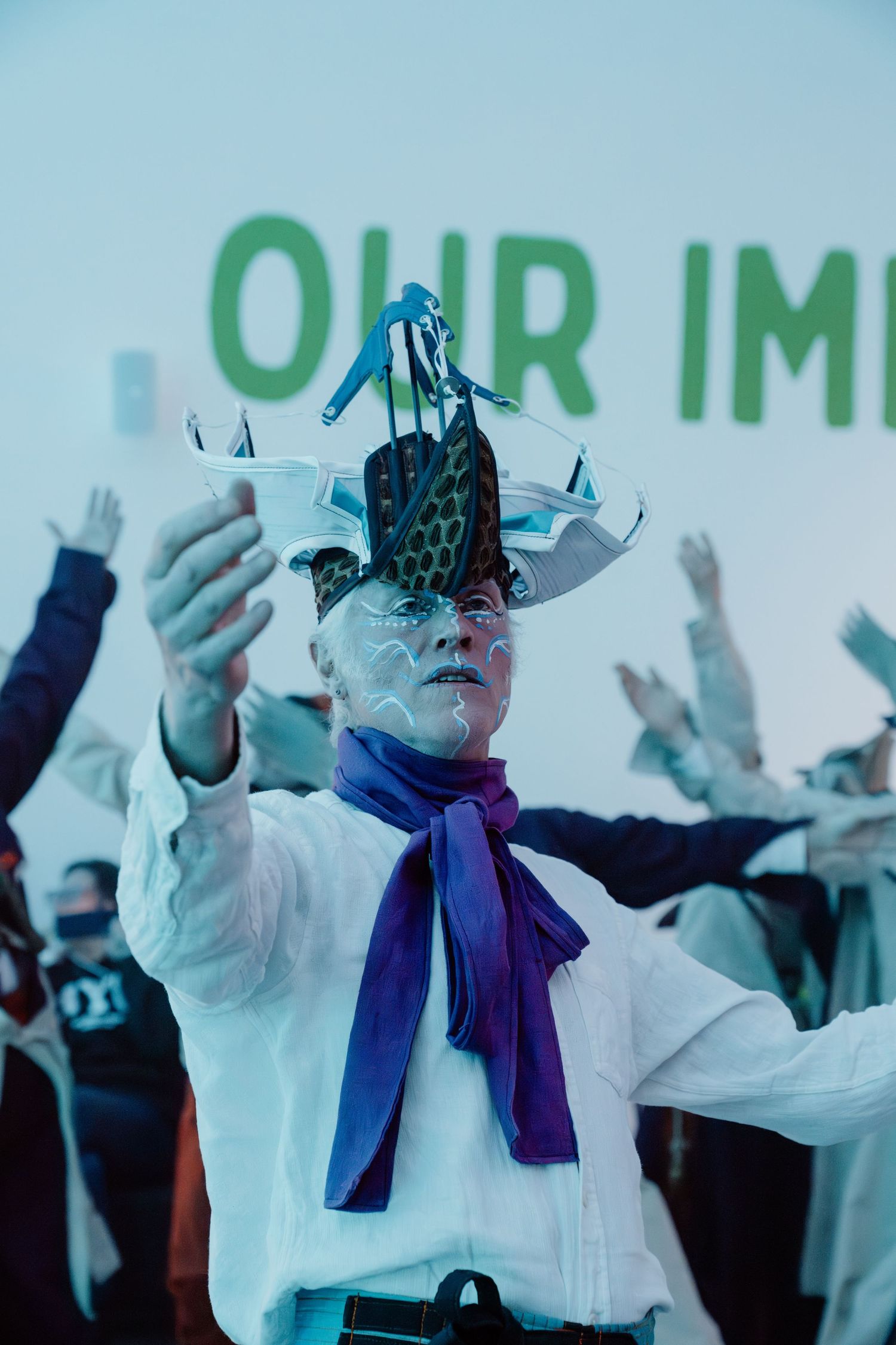 Performer in white costume with elaborate headpiece and face paint gestures on stage before a green projected backdrop.