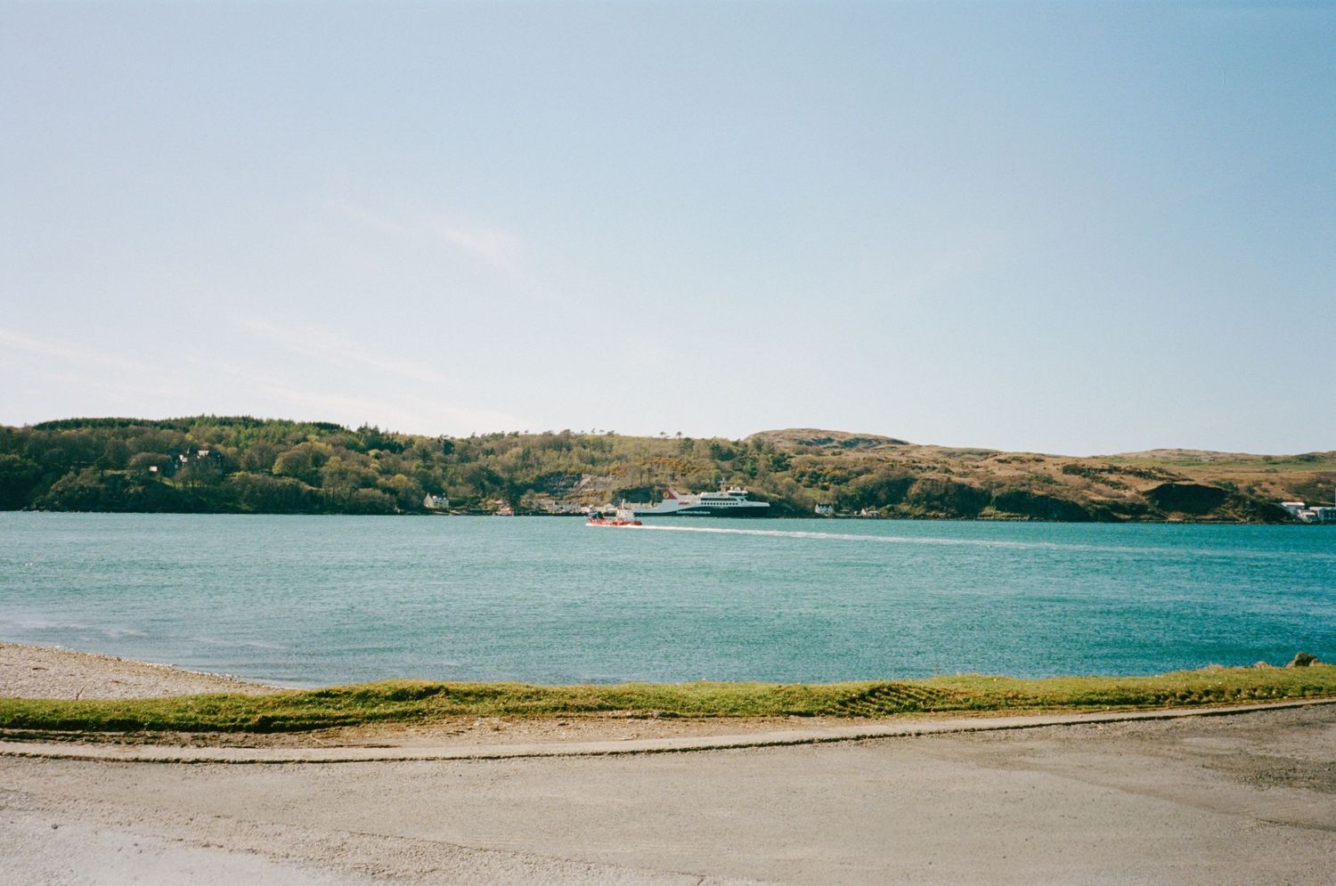 Scenic coastal bay with turquoise water, moored boats, and green hills under a clear blue sky.