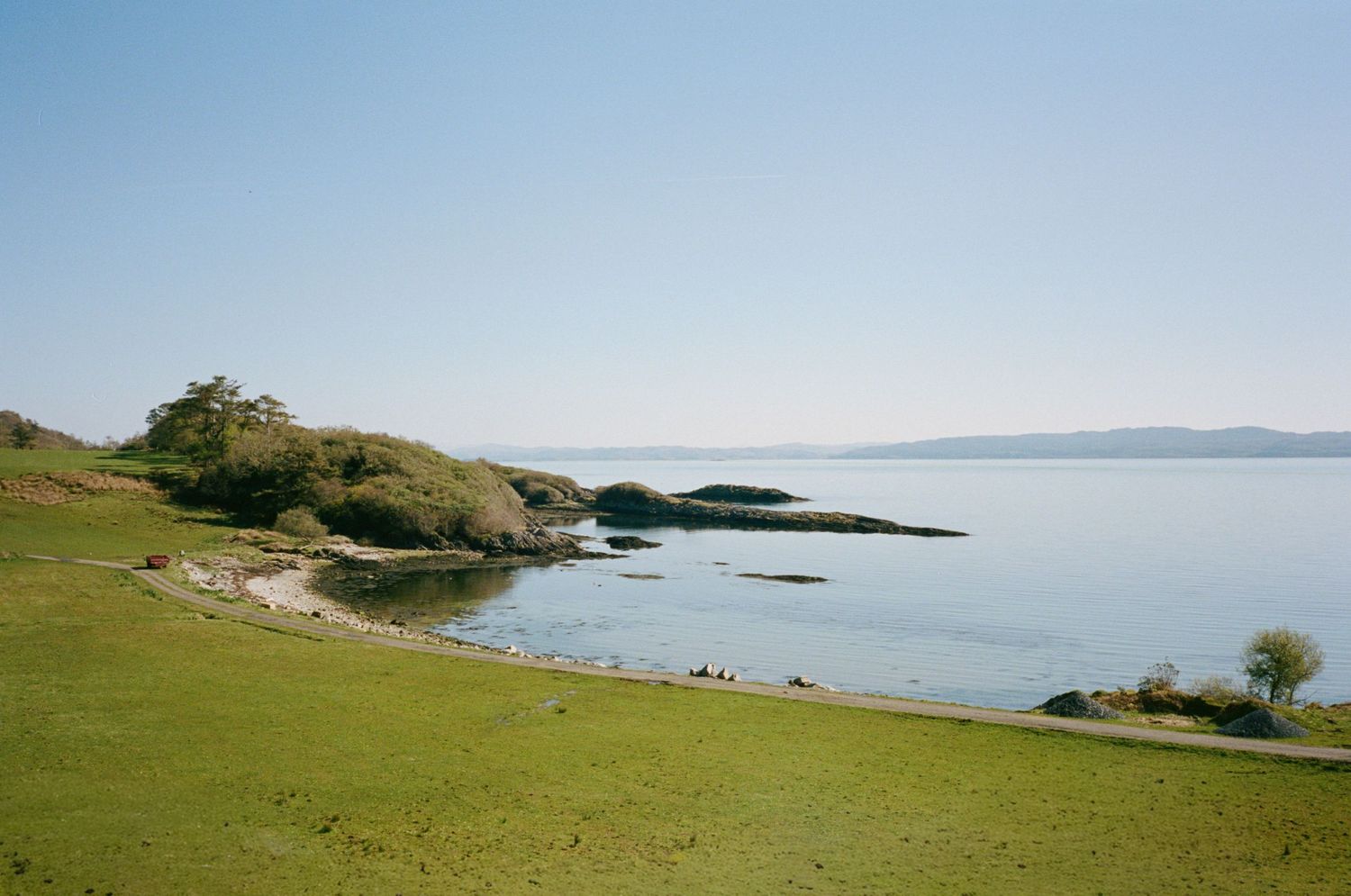 Scenic coastal landscape with green meadows, calm blue waters, and a small rocky peninsula under a clear sky.