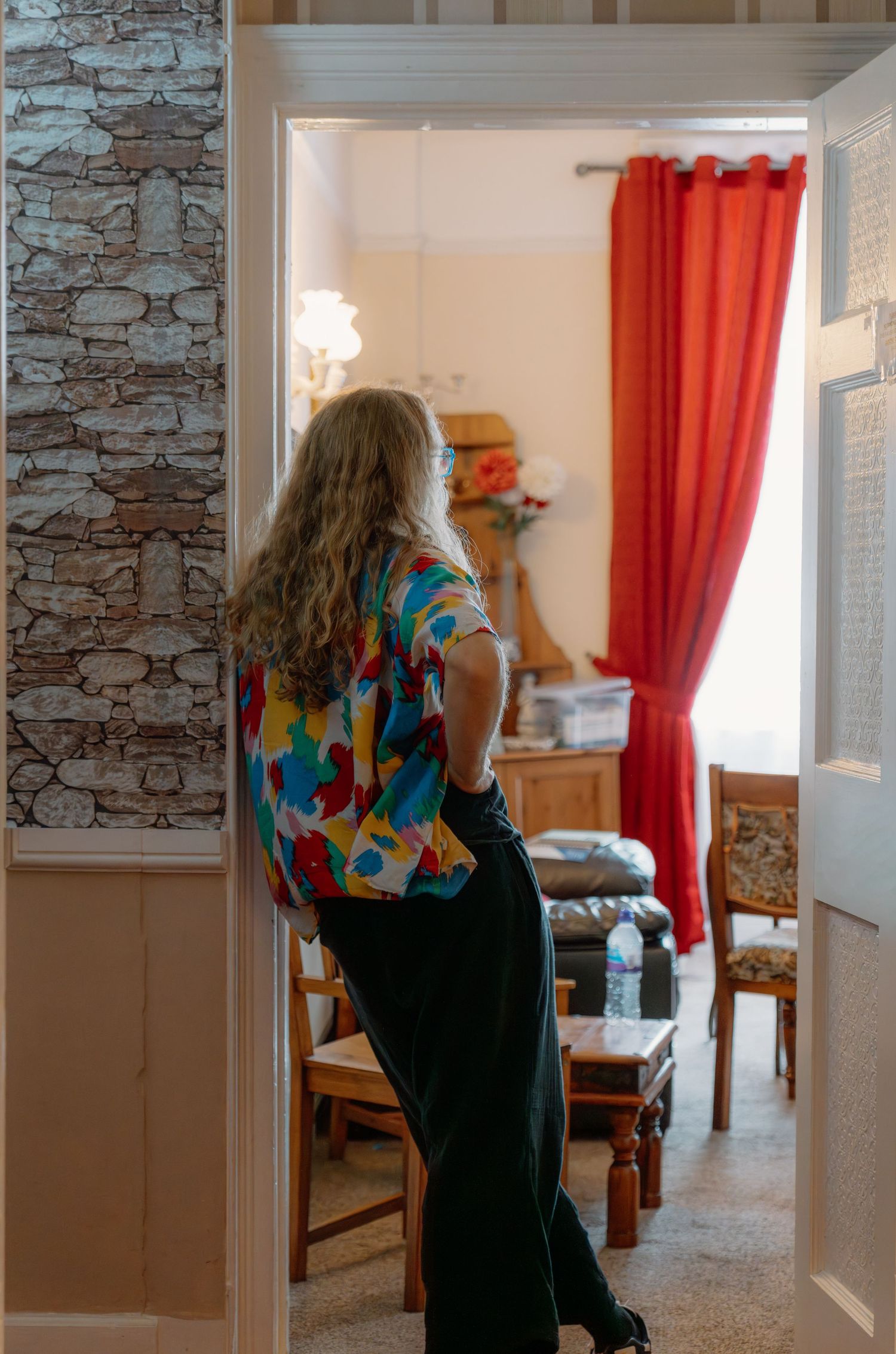 Woman in colorful floral top and black pants standing in doorway looking into room with red curtains.