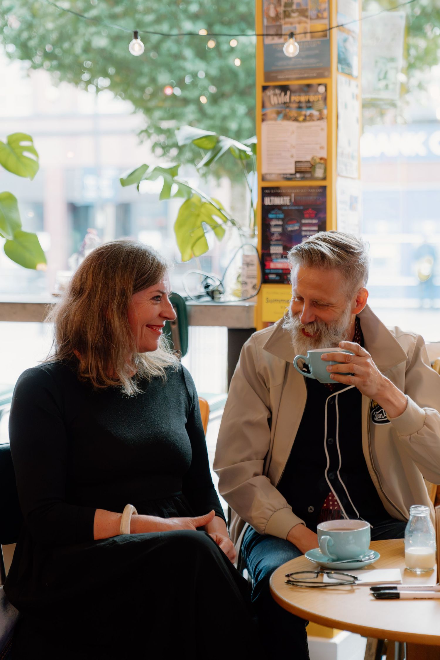 Two people enjoying coffee at a cozy cafe, smiling and conversing in a warm, plant-filled setting.