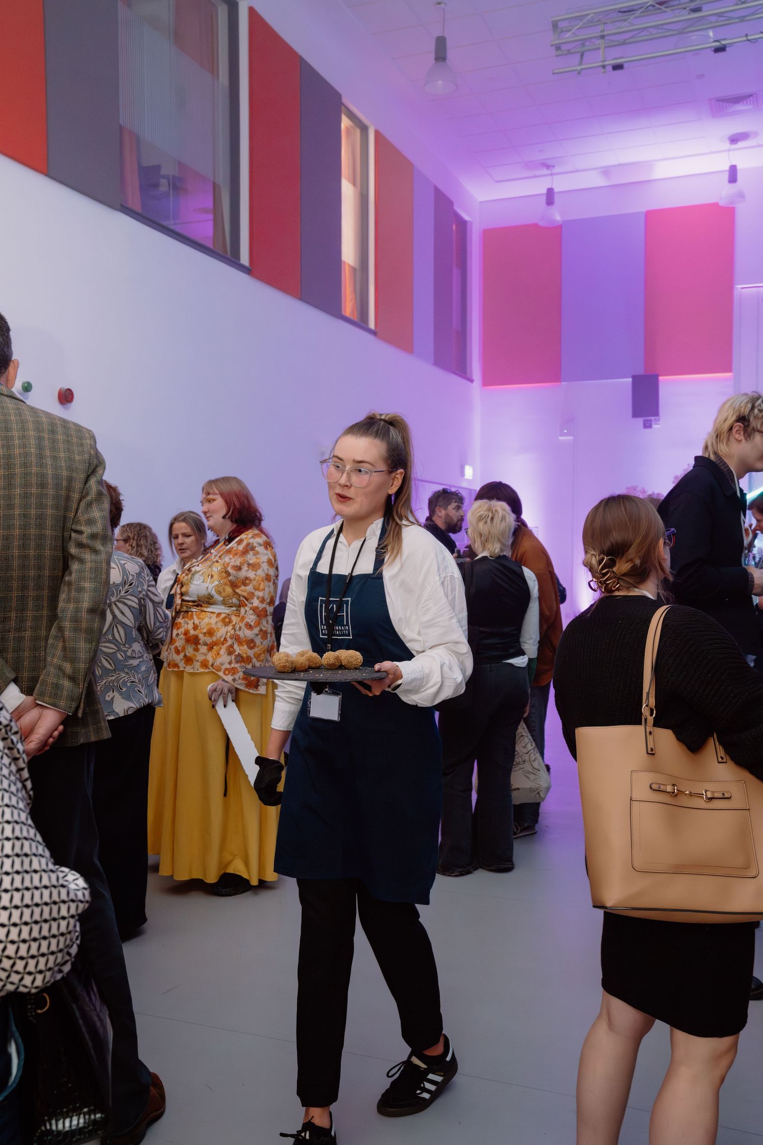 A server in apron holds a tray of pastries at a colorful gallery event with guests mingling in background.