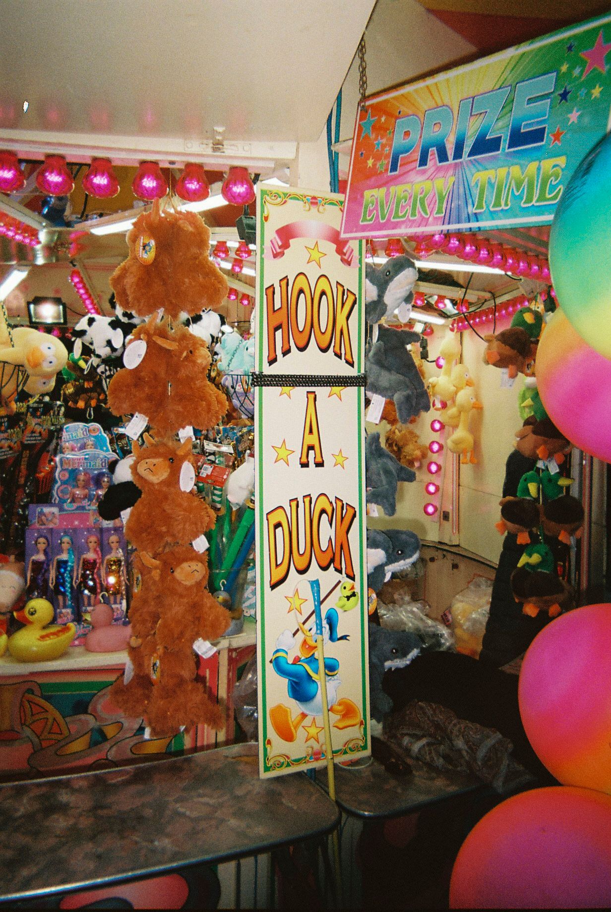 Carnival game booth with colorful balloons, prizes, and signs displaying 'Hook Duck' at a festive fairground setting.