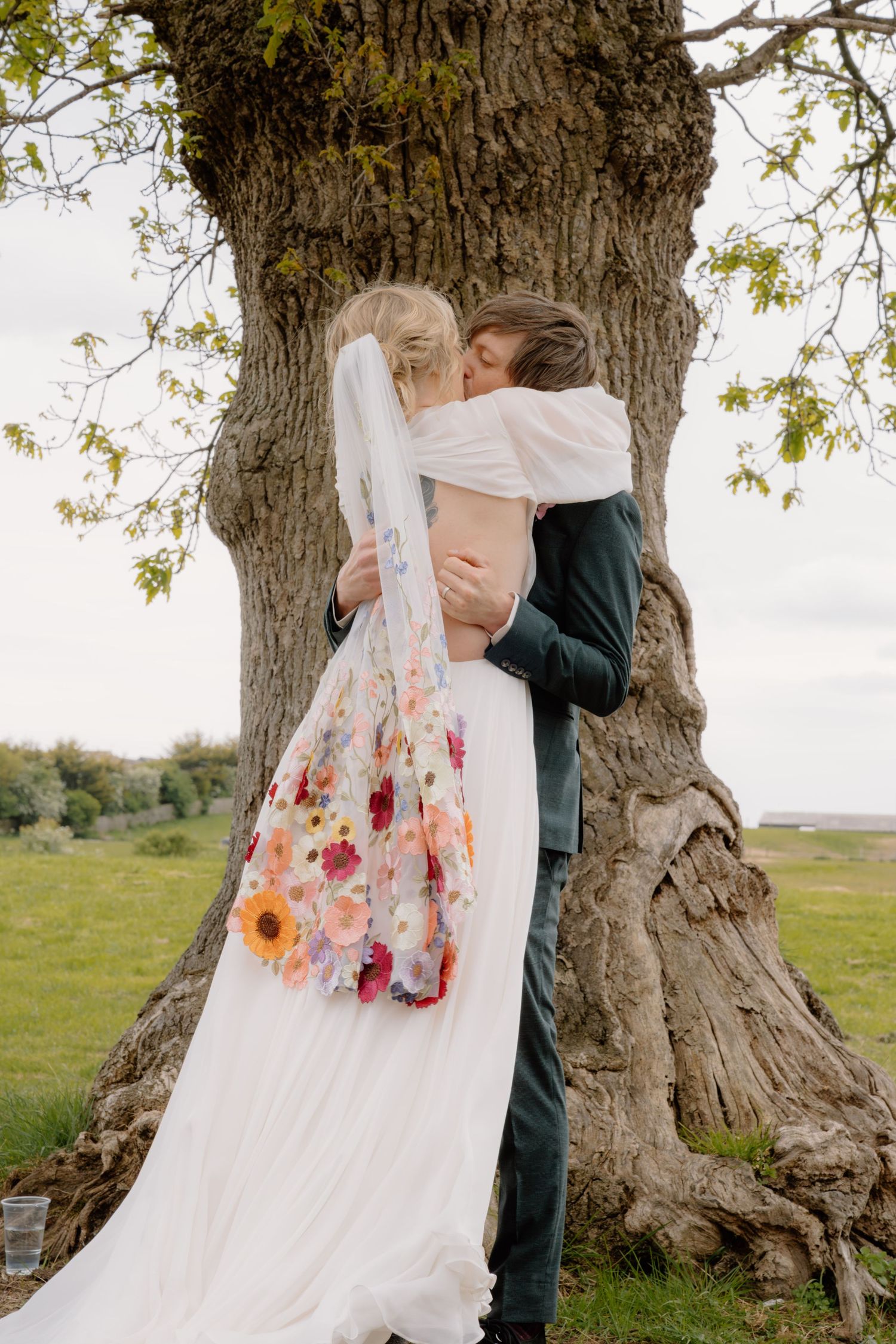 Bride in floral wedding dress and veil kisses groom in dark suit beside large tree in green field.