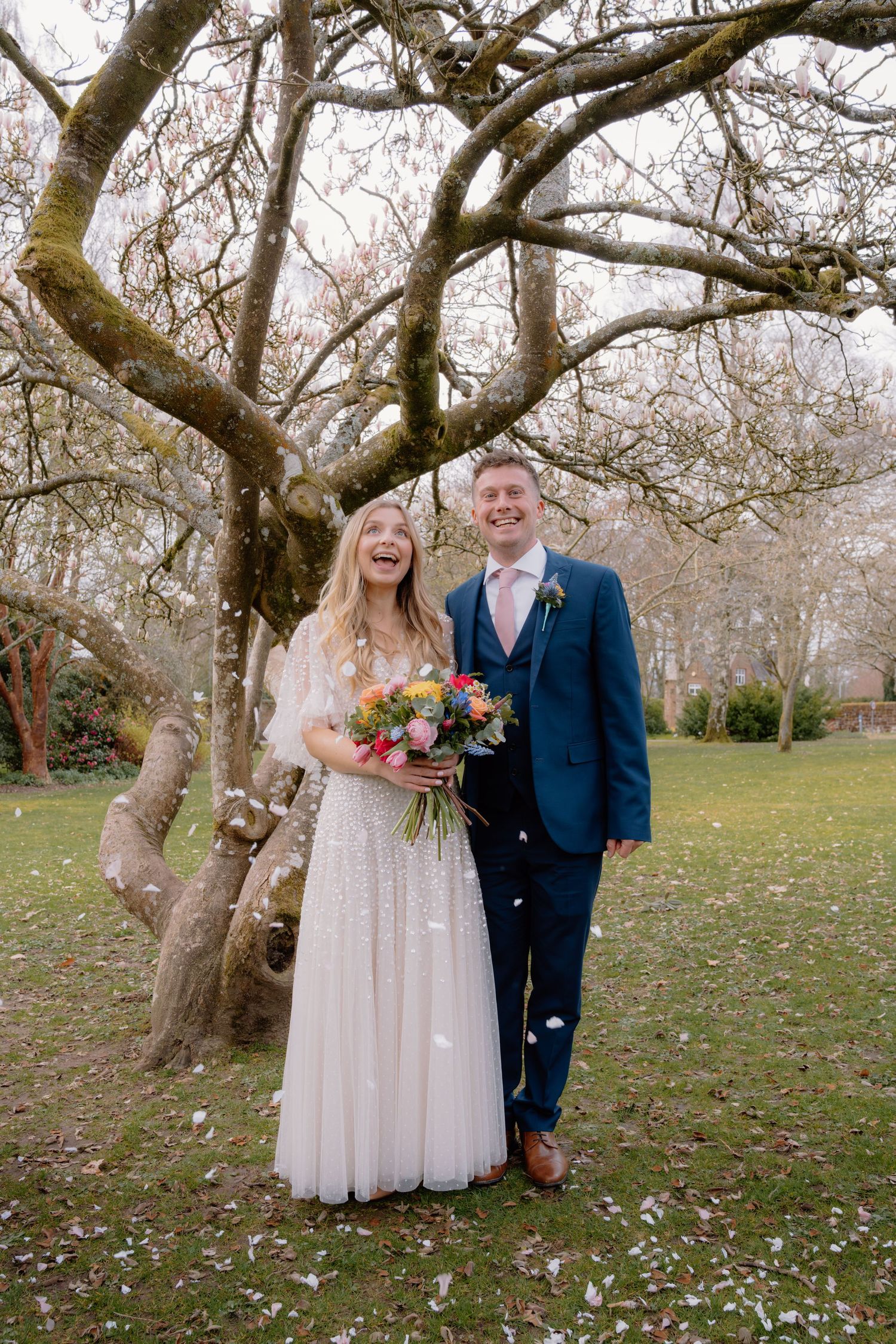 Happy bride and groom laughing under a bare tree, bride holding colorful bouquet, groom in navy suit, outdoor wedding.