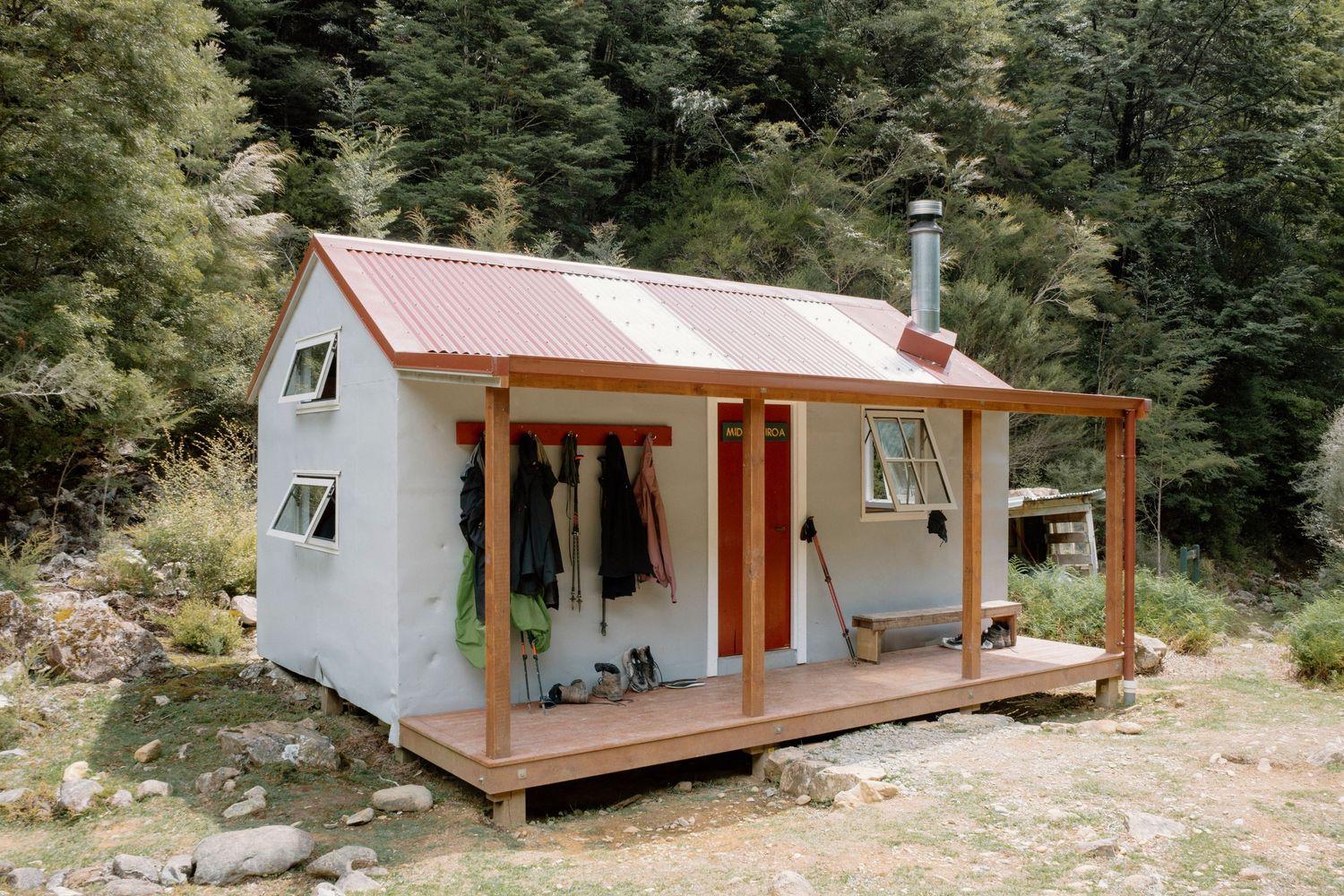 A cozy A-frame cabin with a wooden deck and red metal roof sits nestled among green forest trees.
