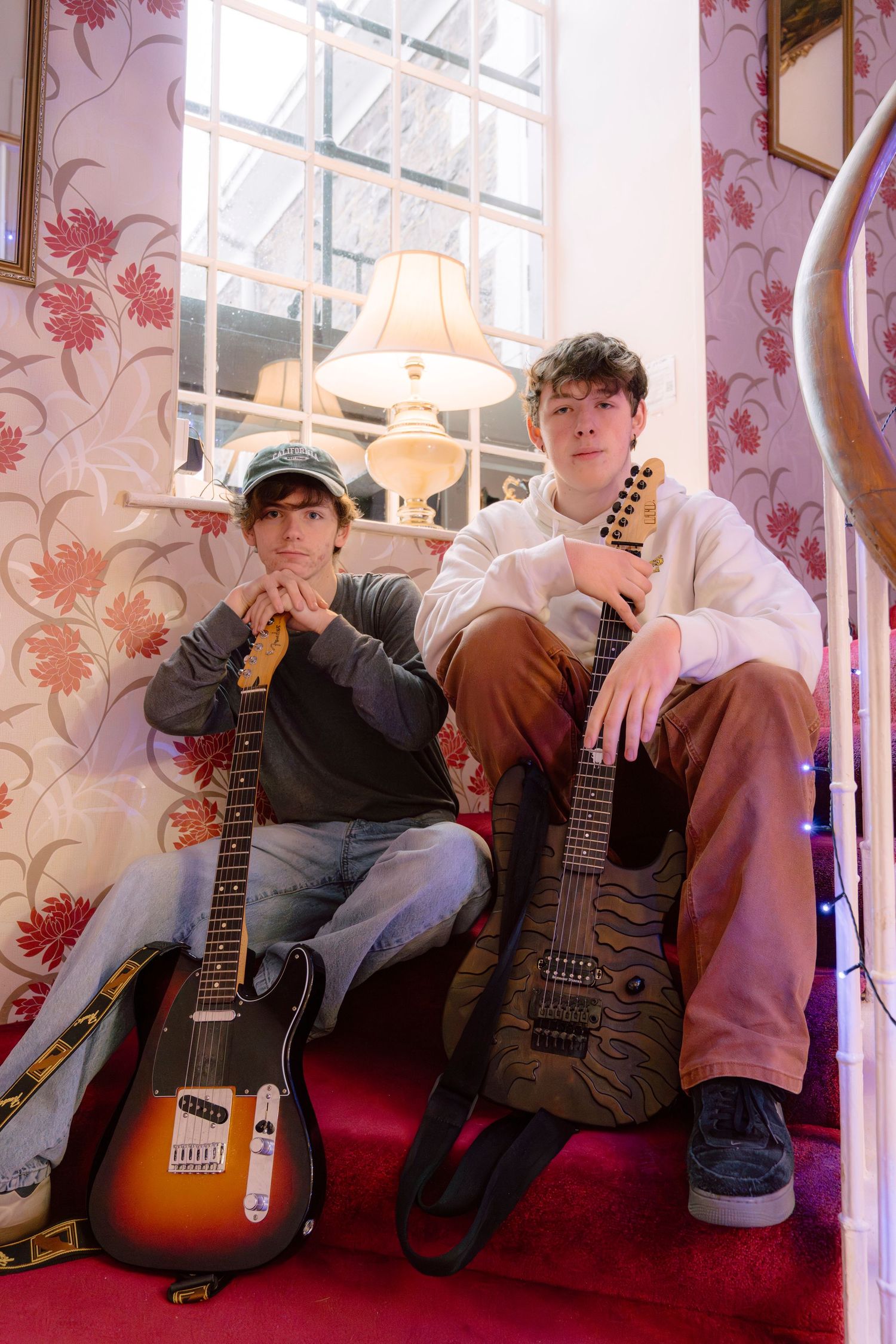 Two young musicians sitting on stairs holding electric guitars in a vintage room with floral wallpaper.