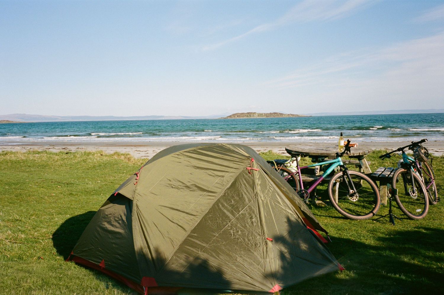 Green camping tent and bicycles on grassy coastal campsite with ocean view and small island in background.