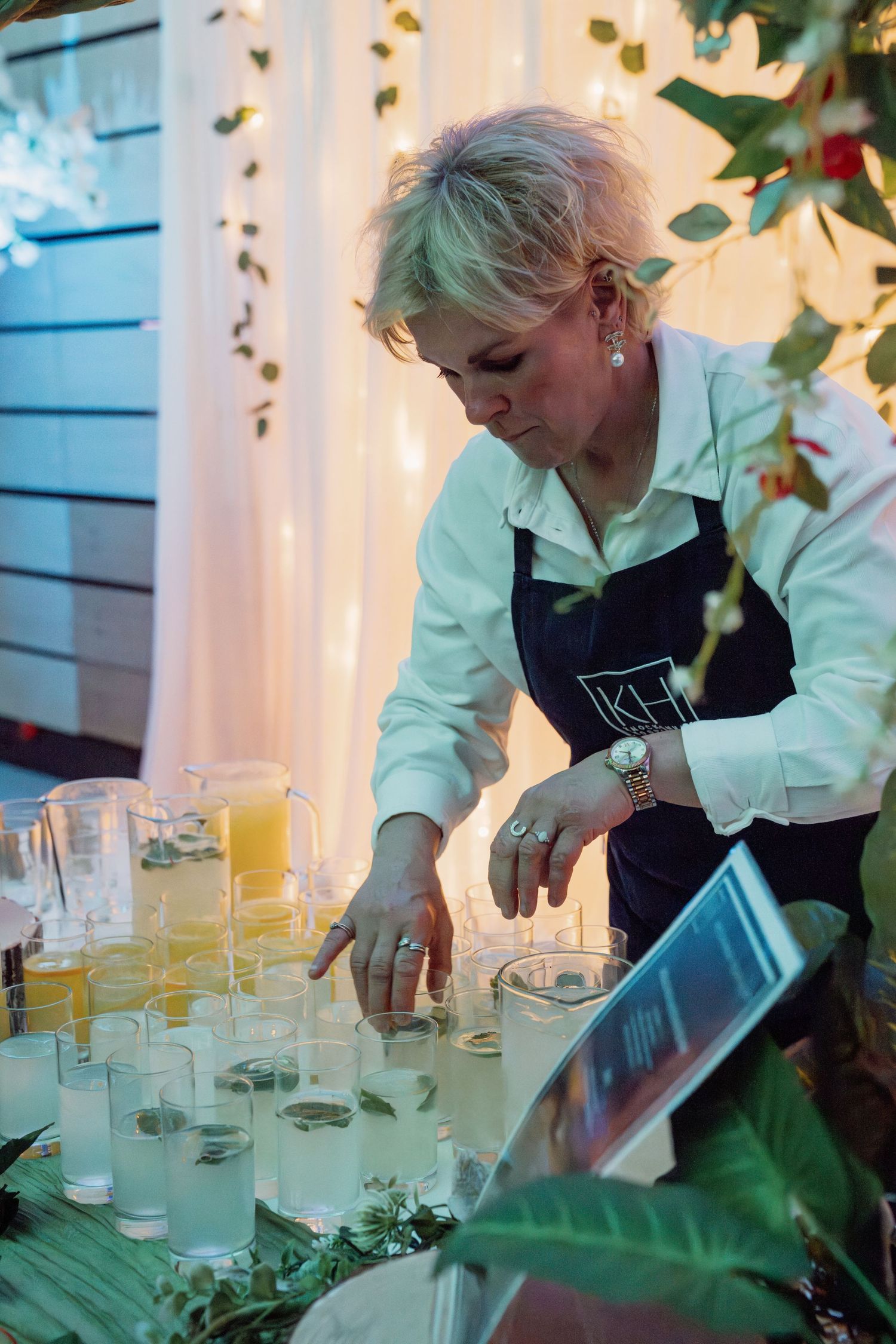 A woman in a black apron arranges glasses on a decorated event bar table with pitchers of juice.