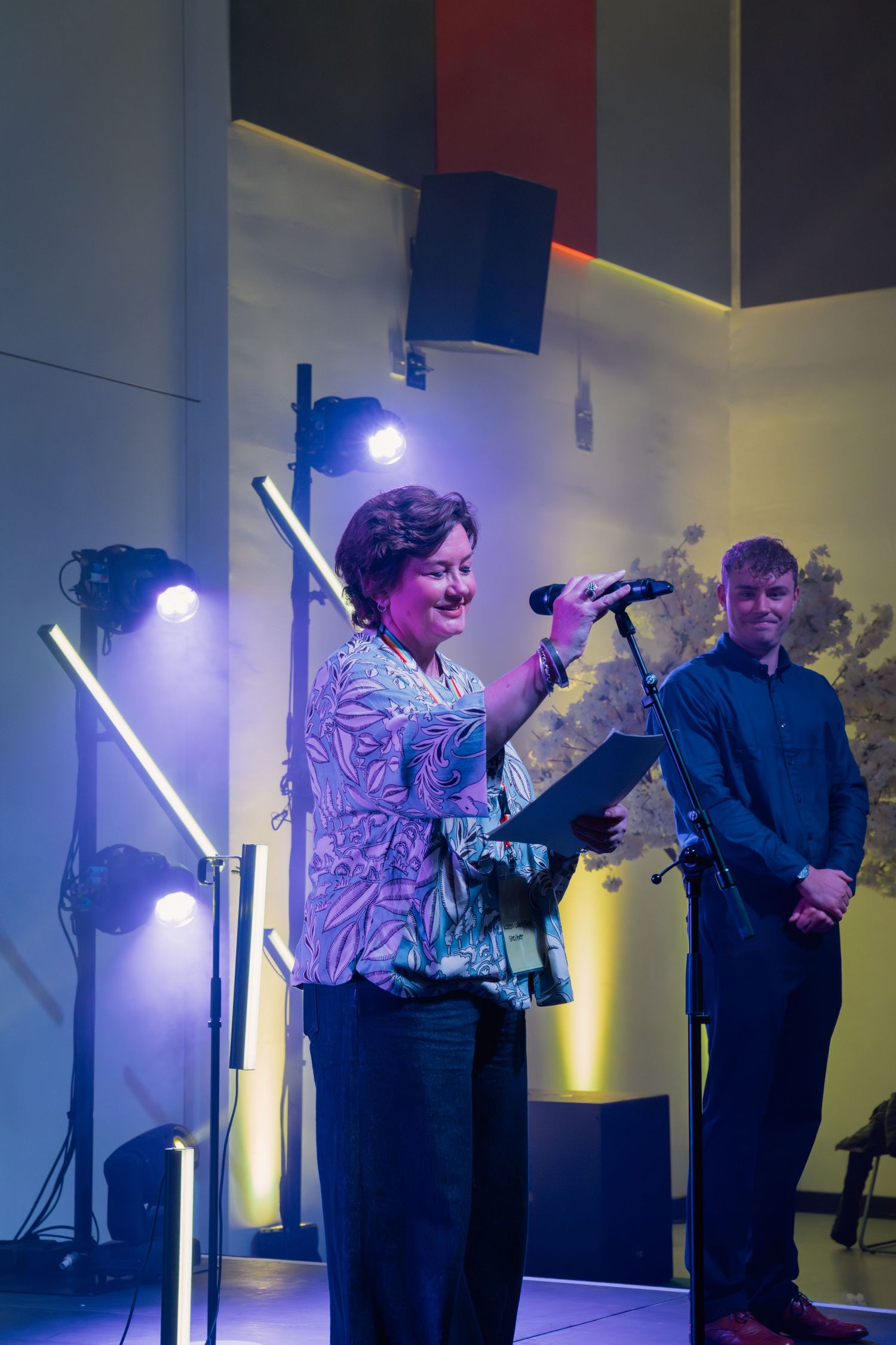 Woman smiling on stage holding microphone and folder at indoor event with colorful stage lighting.
