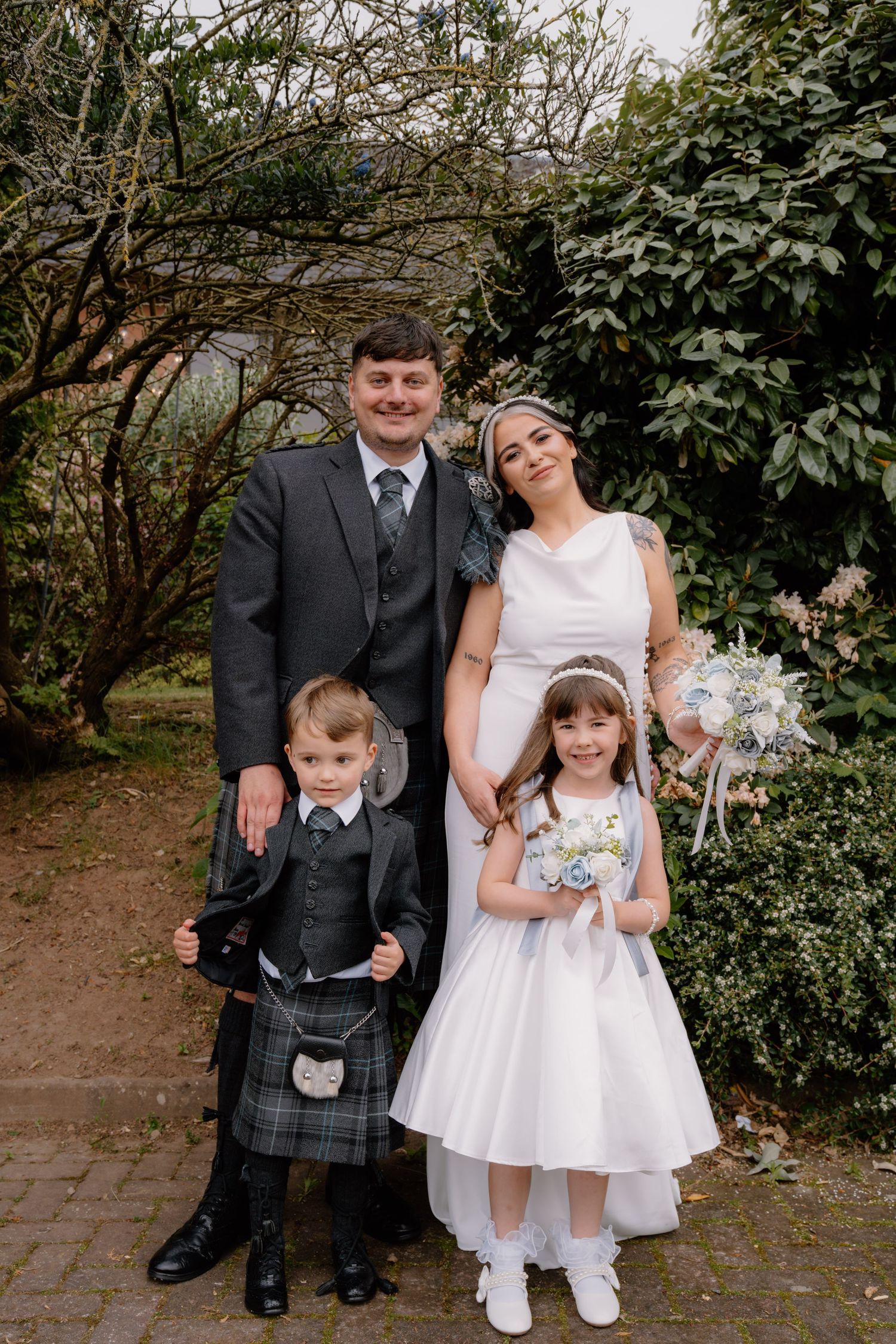 Wedding family portrait with groom in kilt, bride in white gown, flower girl, and ring bearer outdoors.