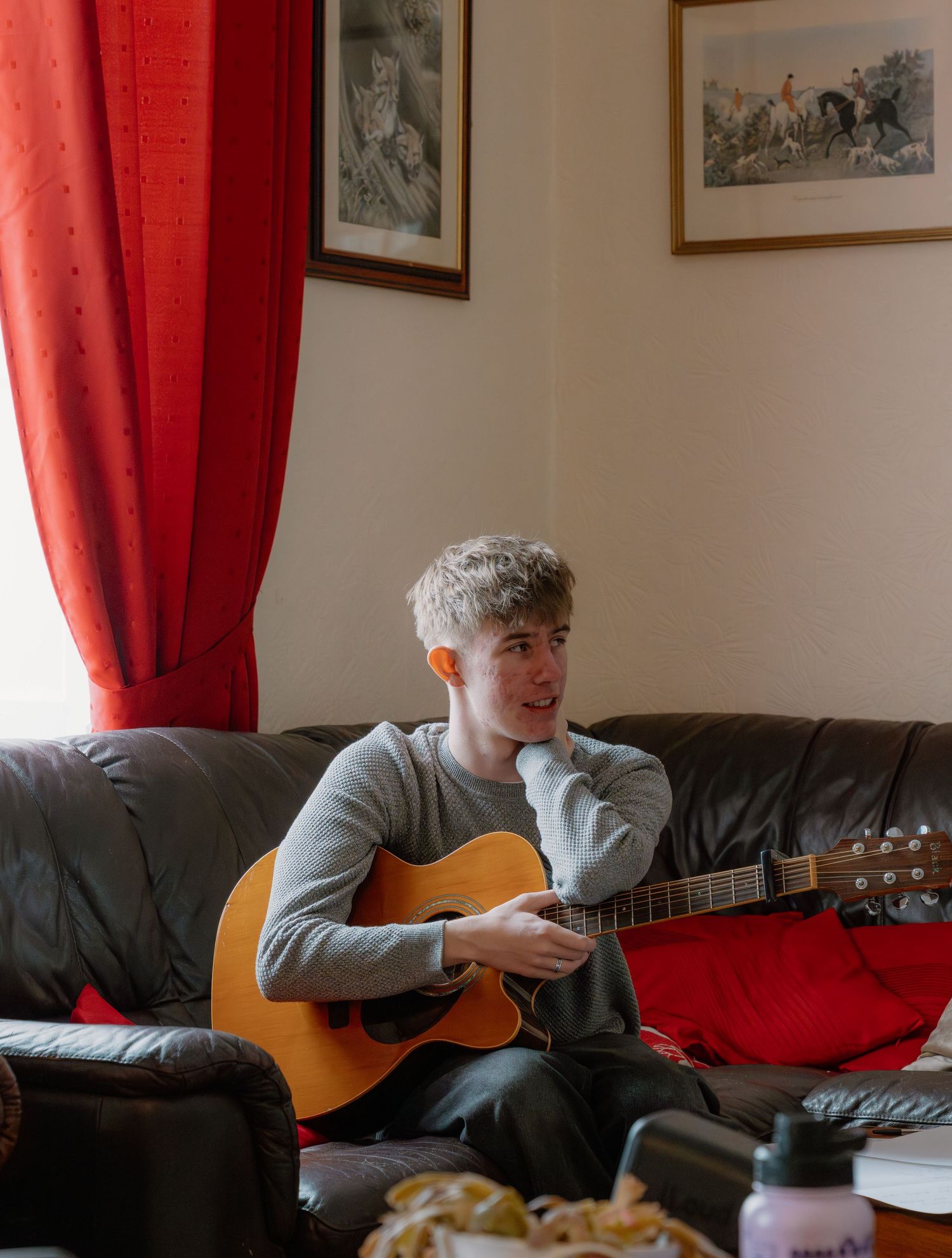 Young man with gray hair playing acoustic guitar on leather couch with red curtains and framed art.