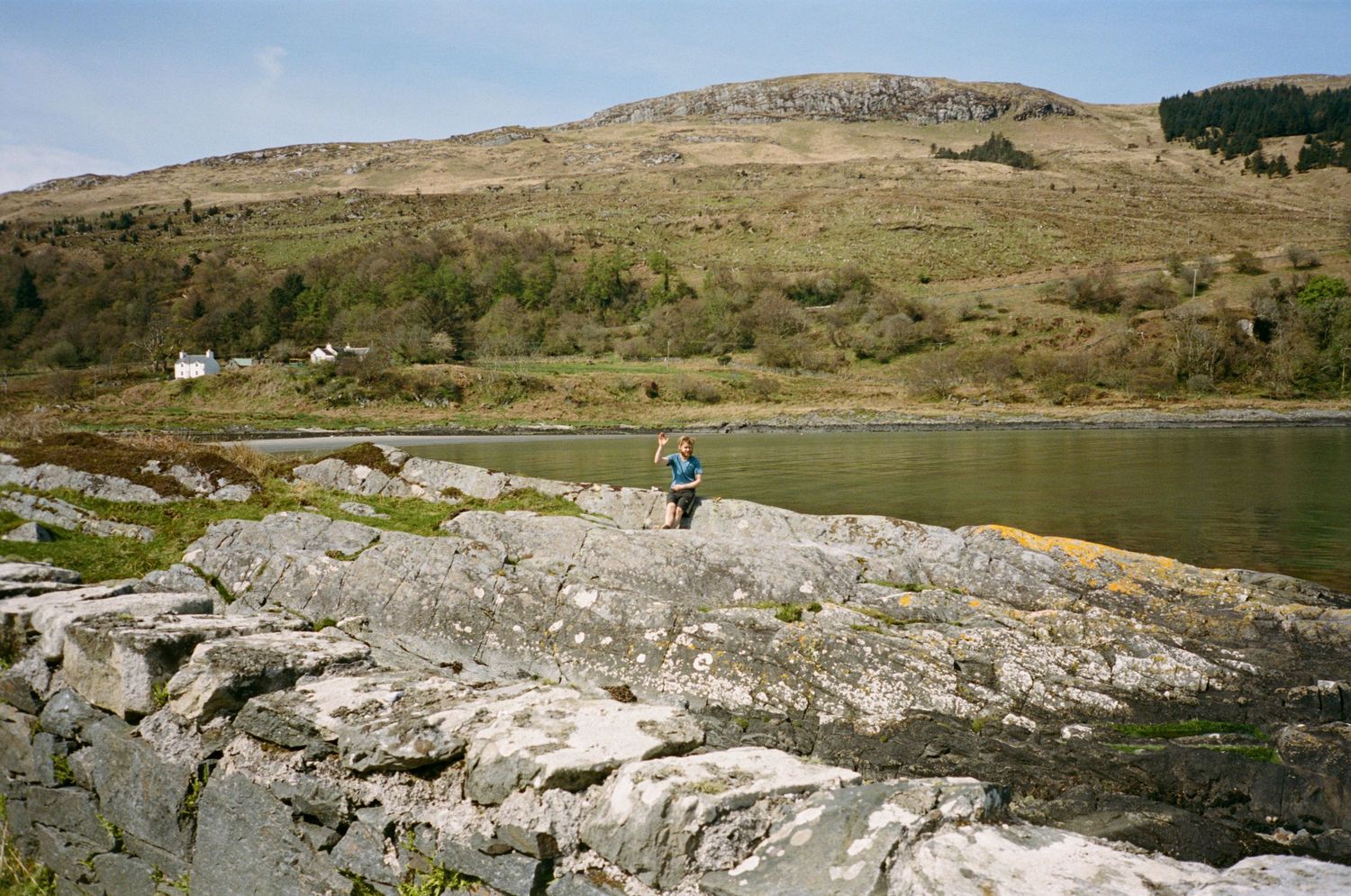 Person standing on rocky shoreline beside a calm loch with green hills and a white cottage in the background.