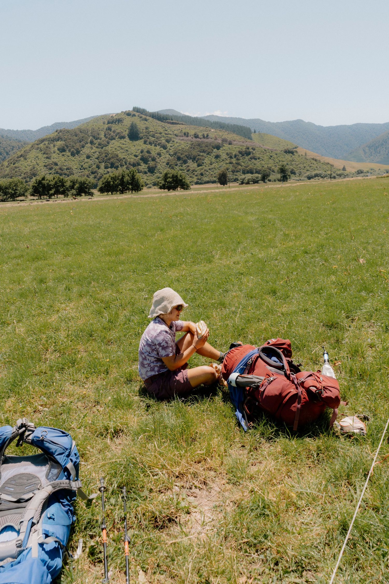A hiker takes a break with backpacking gear on a grassy hillside with mountains in the background.