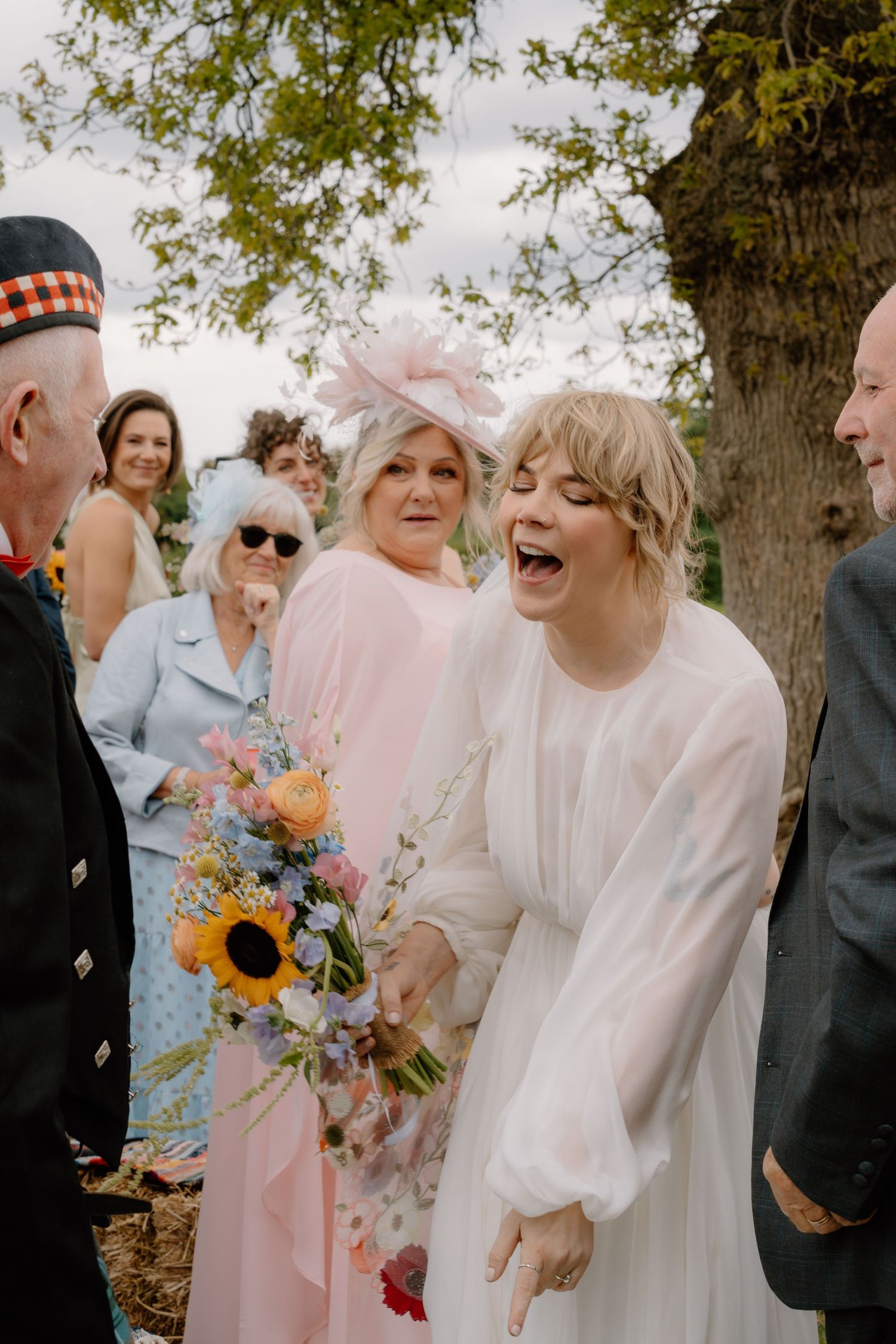Joyful bride in white dress laughing with colorful bouquet including sunflowers surrounded by wedding guests outdoors.