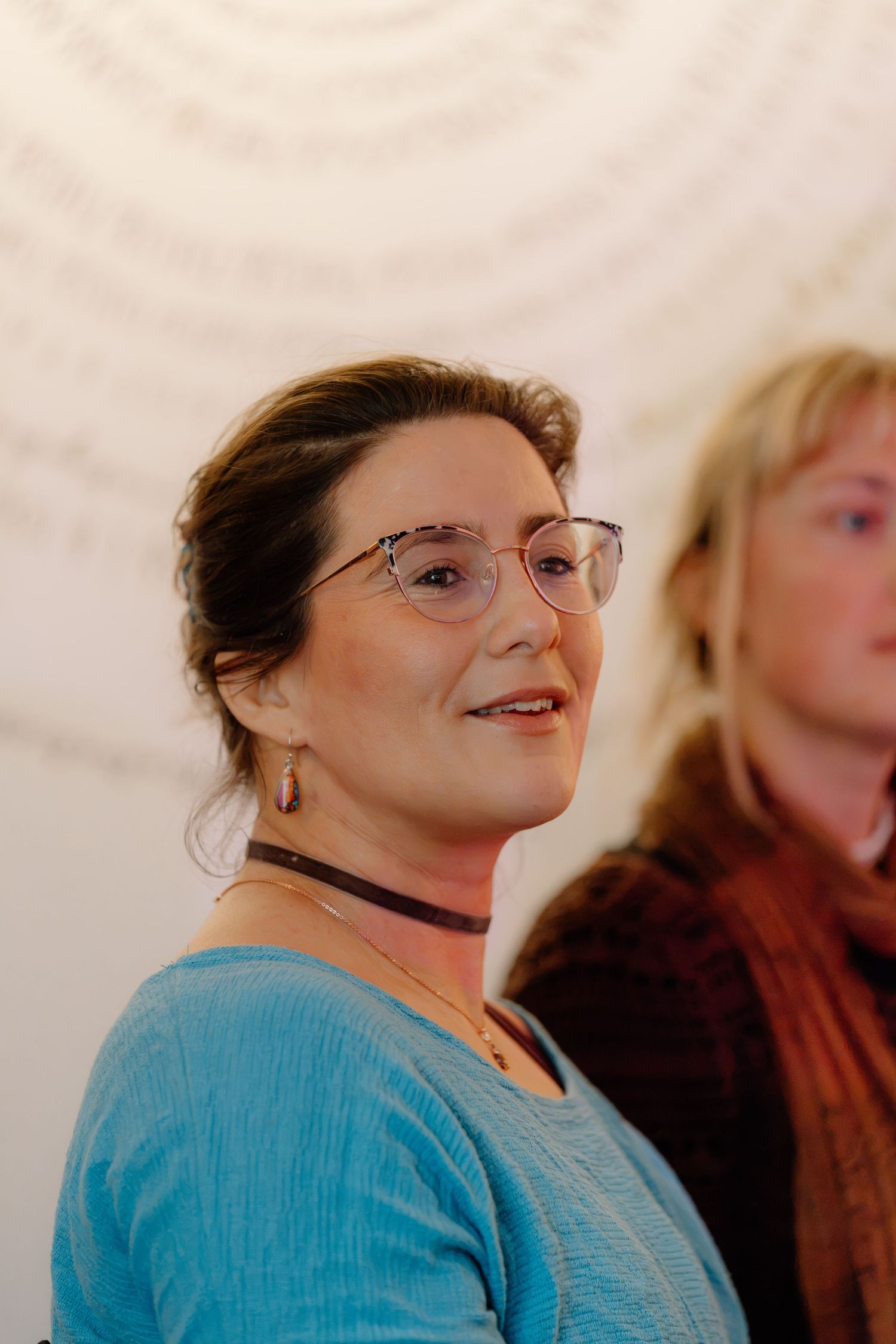 Woman with glasses and blue top smiling, wearing choker necklace, with another person visible in background.