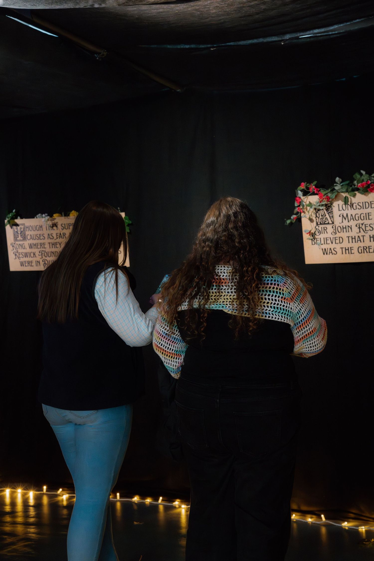 Two women view memorial signs on a dark wall decorated with string lights and holiday greenery.