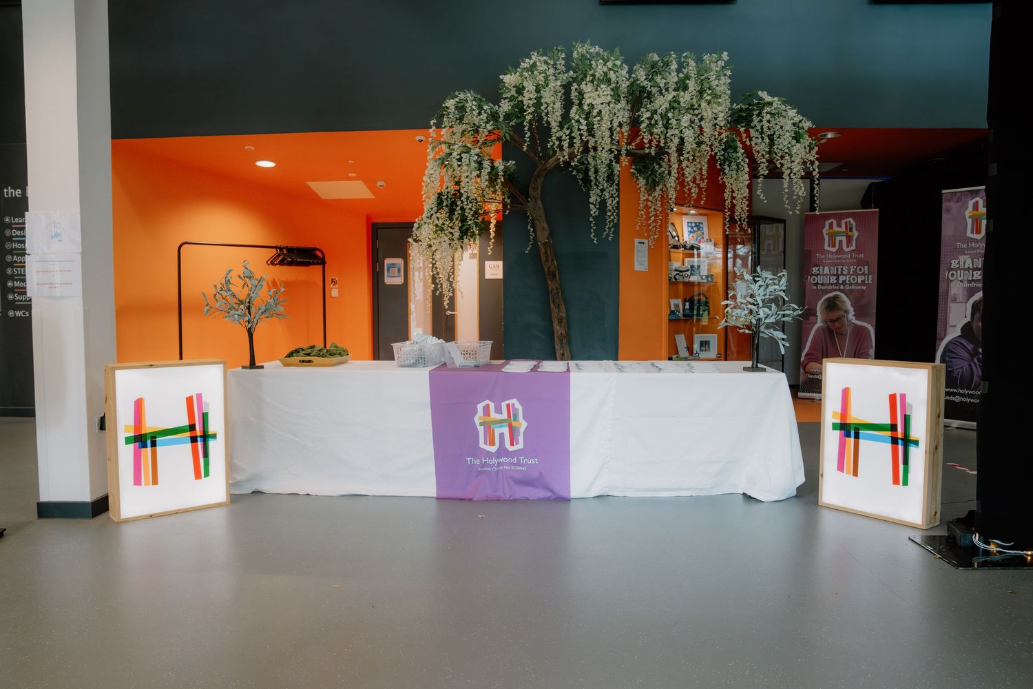 Decorated event table with colorful artwork, white tablecloth, purple logo banner, and hanging wisteria tree in modern venue.
