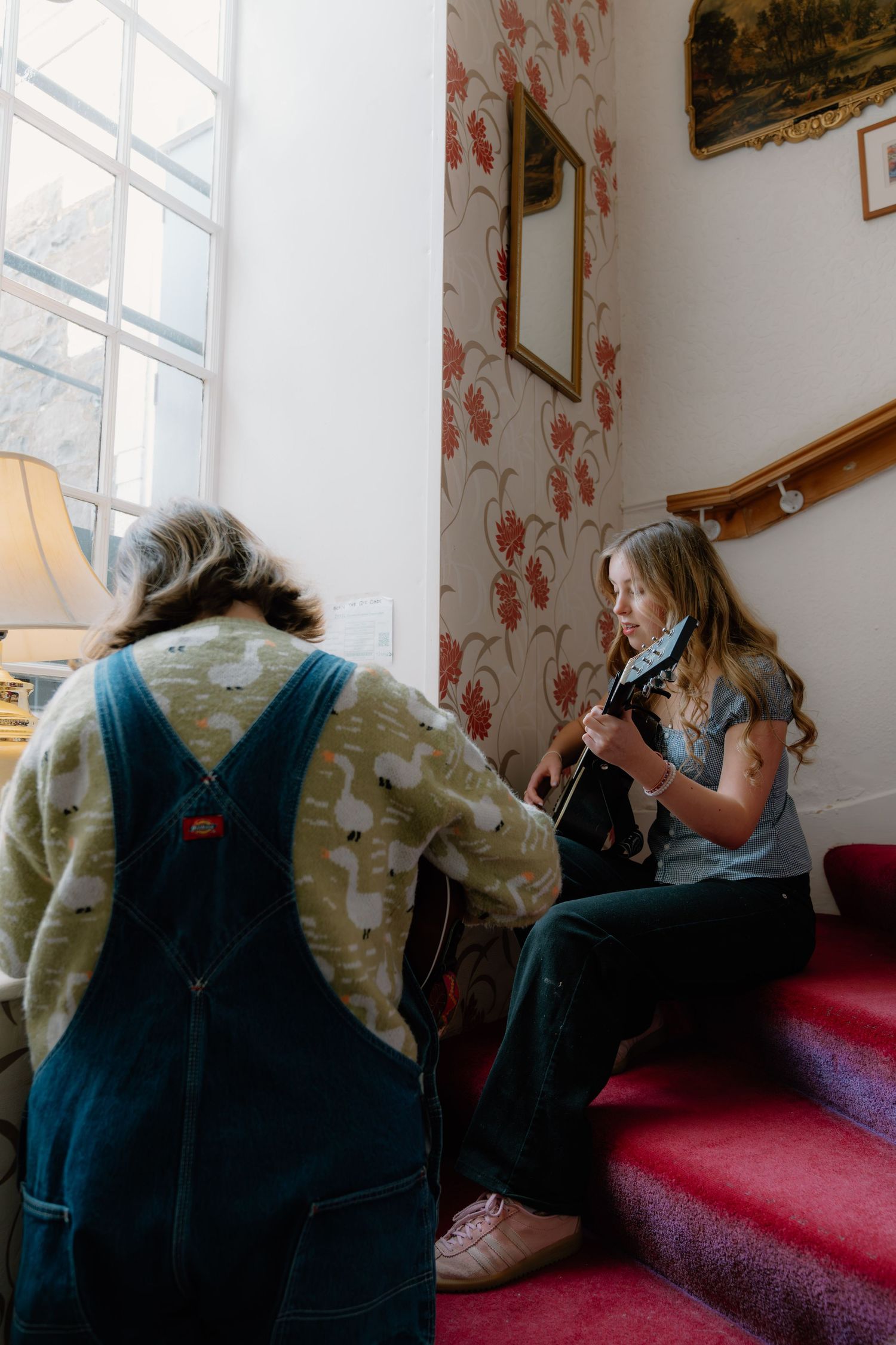 A girl plays acoustic guitar on red stairs while another child in overalls watches in a cozy floral wallpapered room.