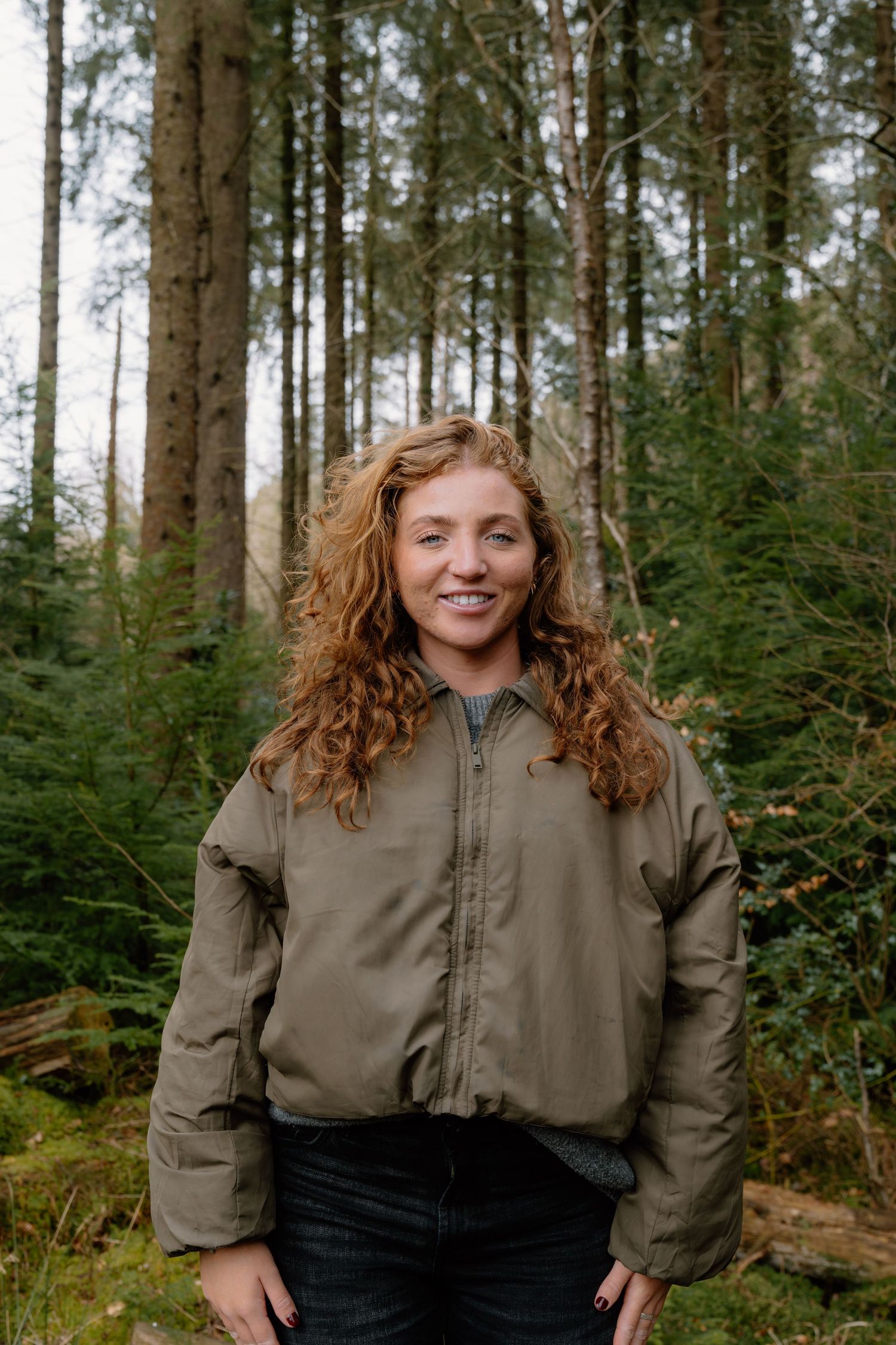 A smiling woman with curly red hair wearing a khaki jacket stands in a lush green forest.