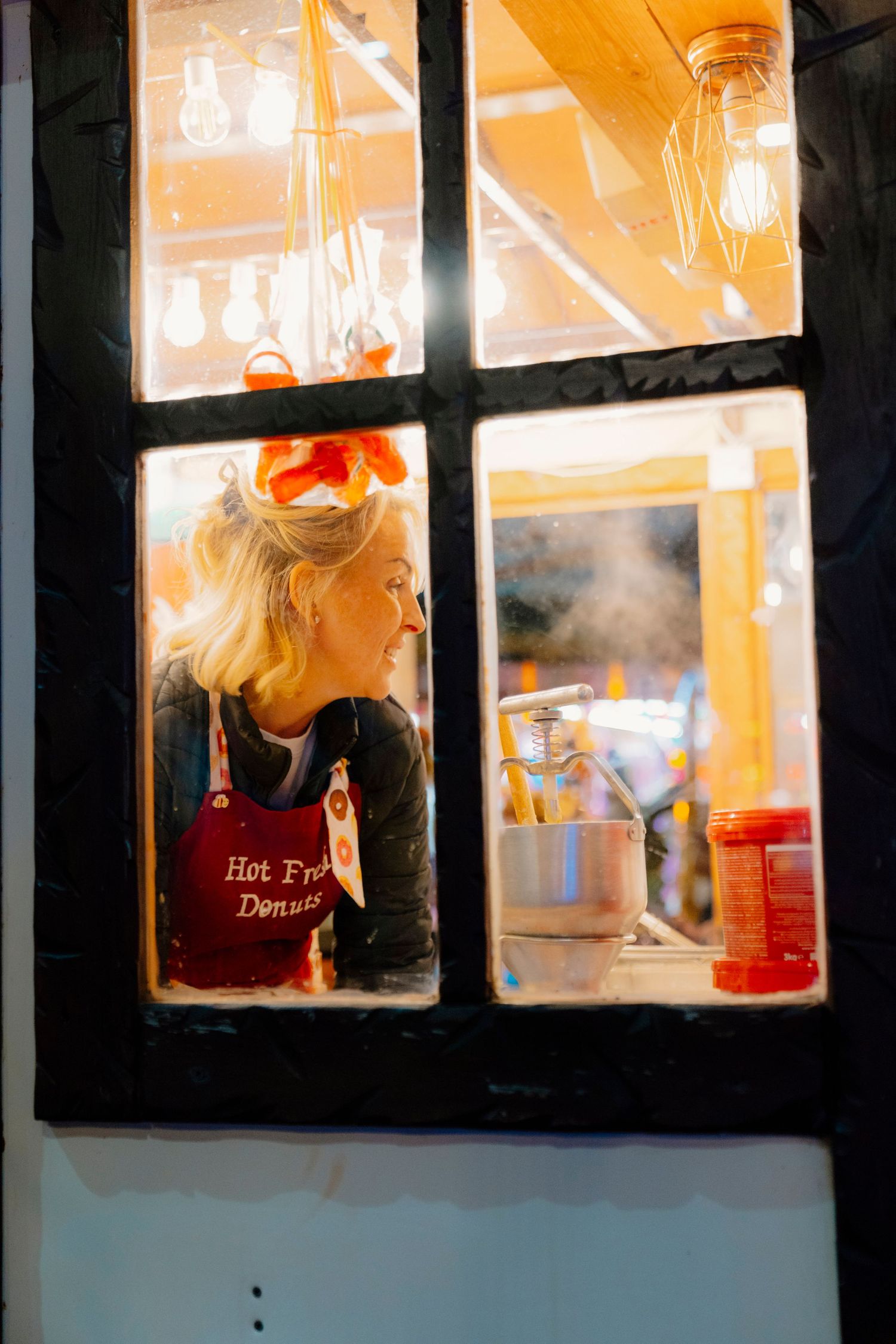 Through a window pane at night, a person in a red apron works in a warmly lit kitchen with steam rising from cookware.