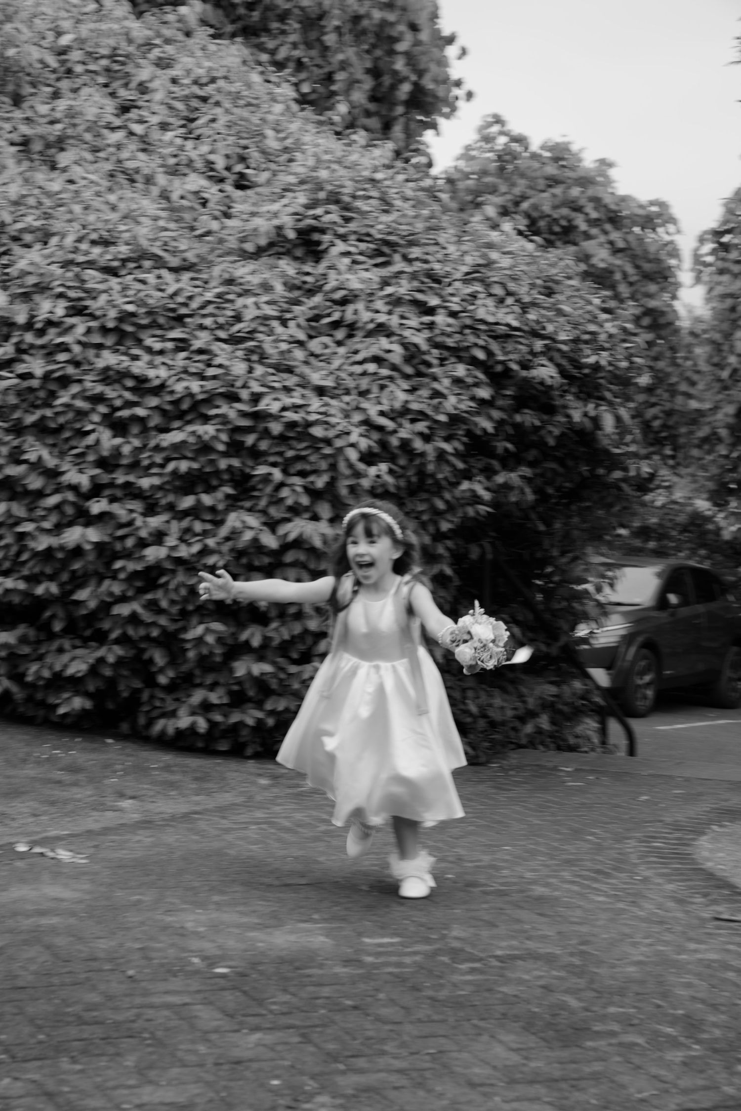 Black and white photo of a joyful little flower girl in a white dress running with a bouquet outdoors.